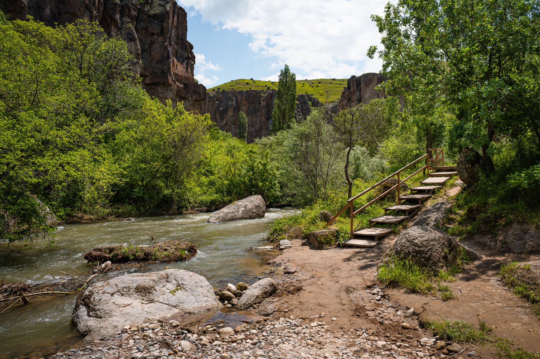 A wooden restaurant platform extending over the Melendiz River in the middle of Ihlara gorge, with canyon walls rising on both sides