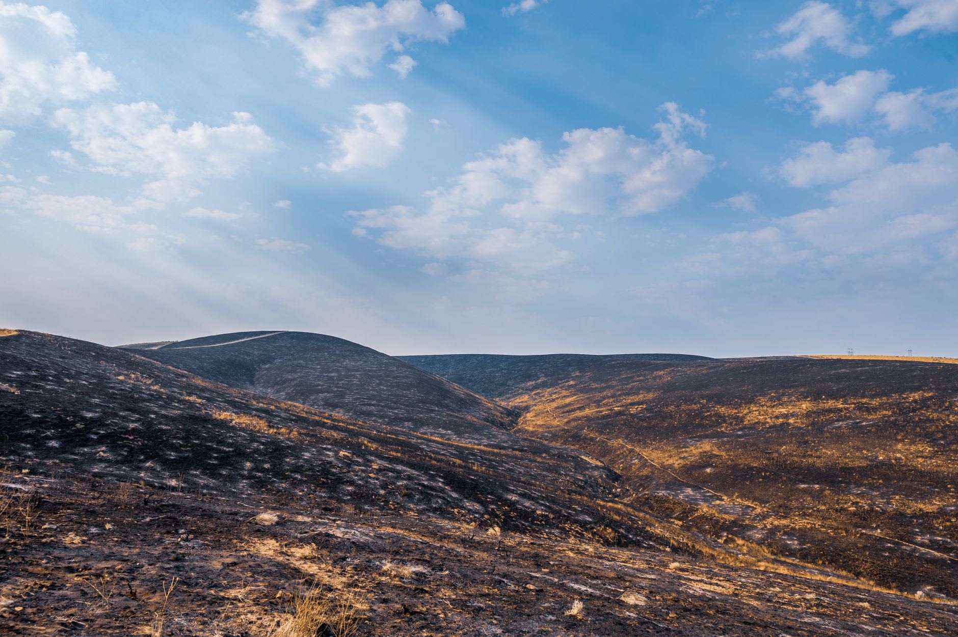 A wall of low orange flames burning along the base of a bare hillside, with a few visitors standing back from the heat at dusk near Baku.