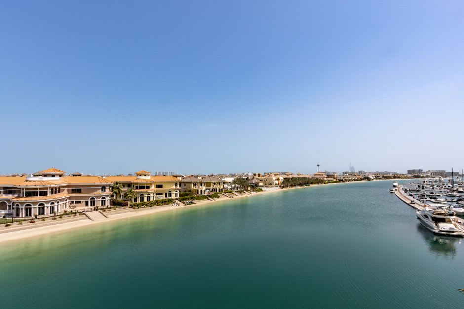 View across a still canal in Amwaj Islands with waterfront villas and a moored boat under a pastel sunset sky