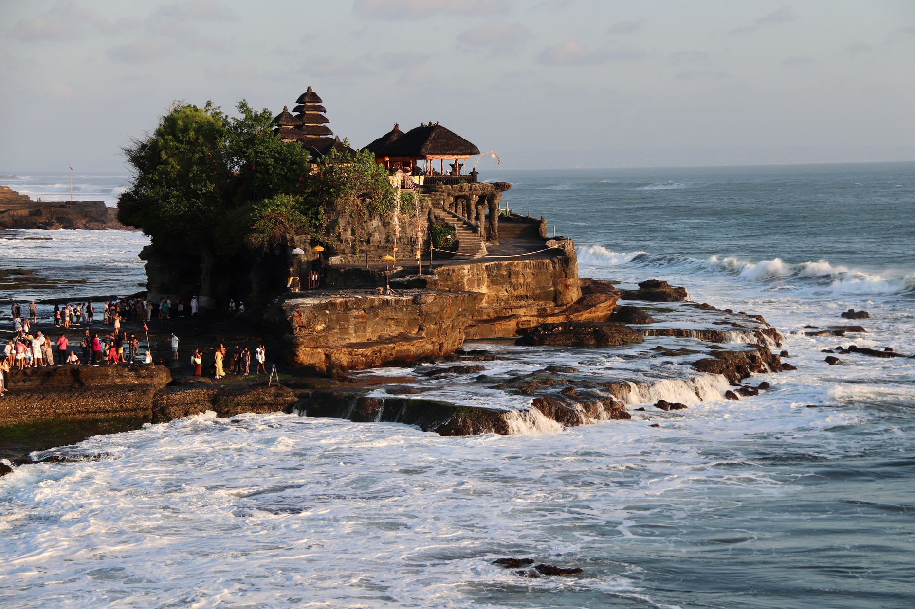 Waves breaking around the base of Tanah Lot's sacred black rock in the morning light, spray rising toward the temple