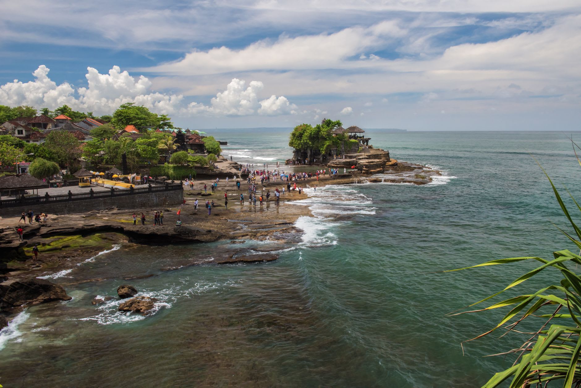 Pilgrims crossing the rock platform at low tide toward Pura Tanah Lot, the temple gate open above the sea