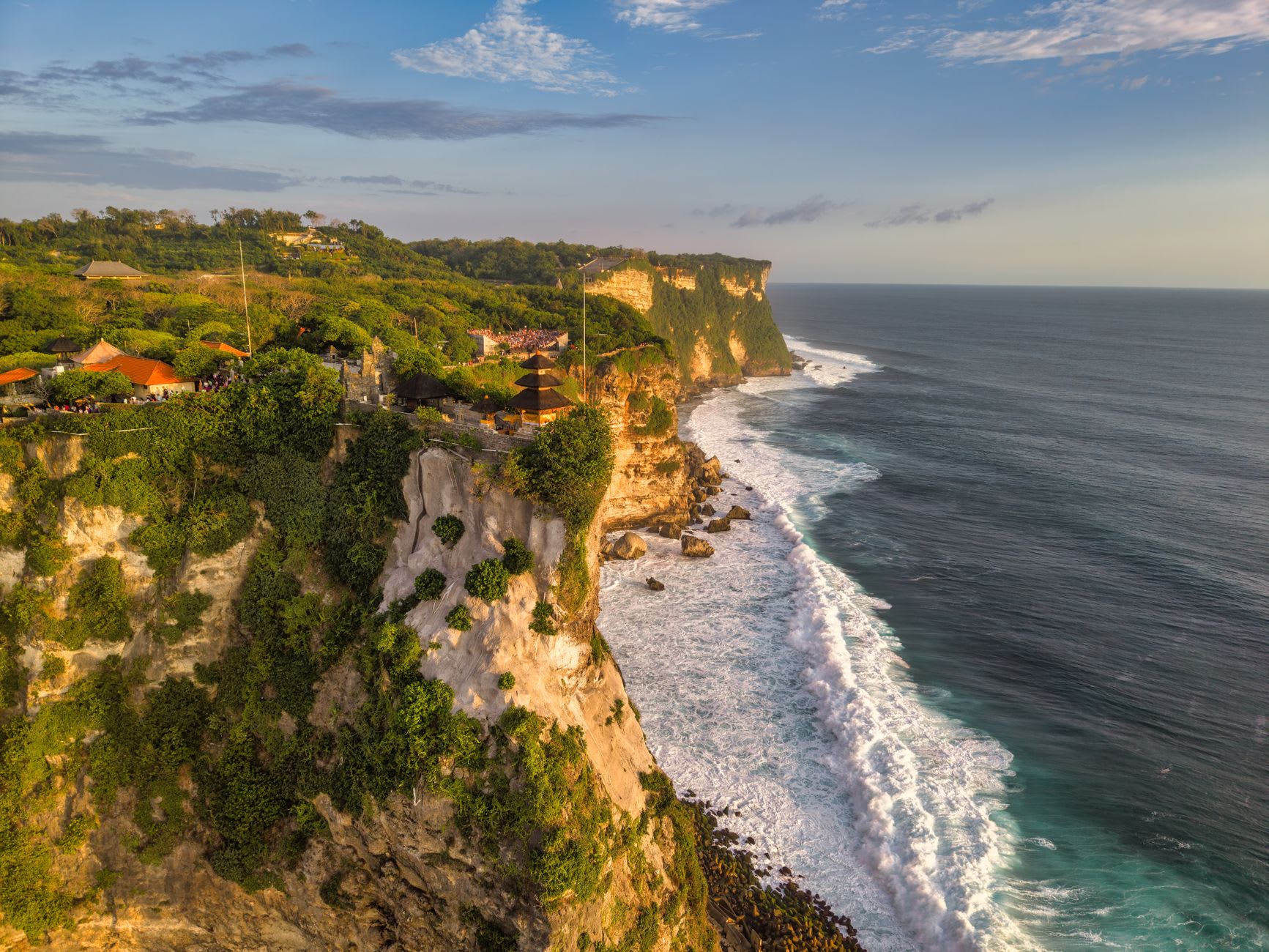 Surfers in the lineup at Uluwatu's main break below the limestone cliff, perfect left-hand wave peeling across the reef