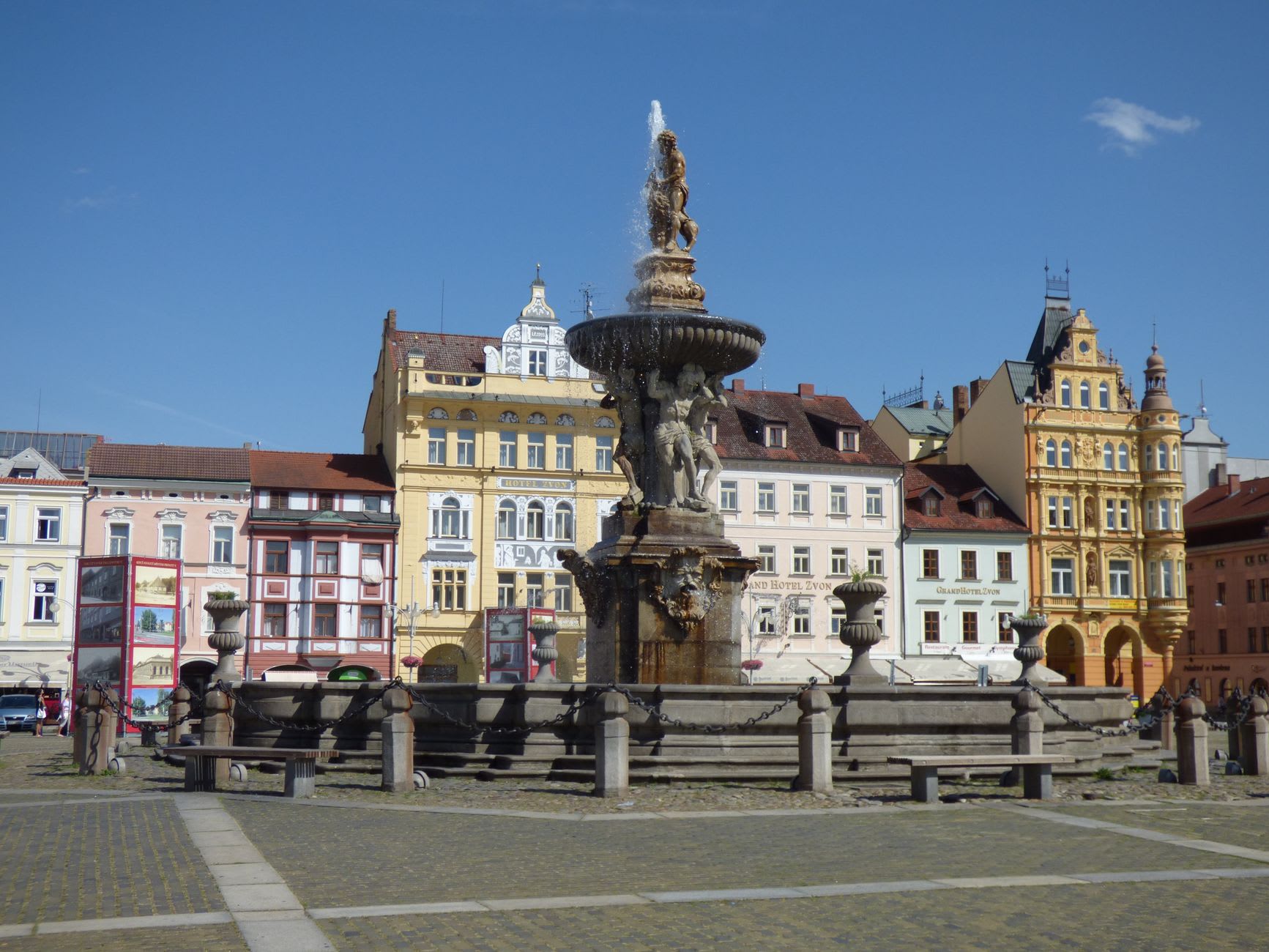 Náměstí Přemysla Otakara II at evening light, the facades catching the last sun, the Samson Fountain silhouetted in the foreground, locals sitting on benches around the square
