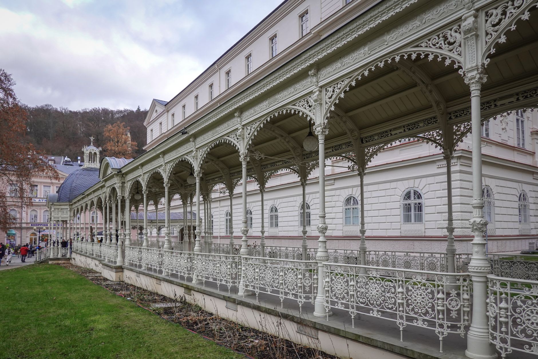 La ferronnerie en fonte blanche et verte de la colonnade principale de Mariánské Lázně dans la lumière du matin, avec la forêt qui monte abruptement derrière