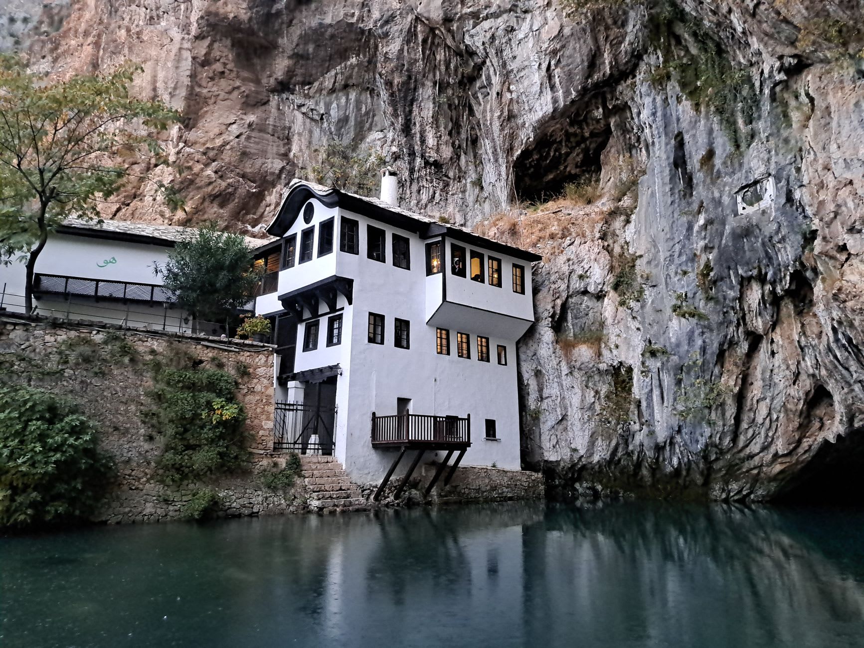 The cool green waters of the Buna source surging from the cliff face, the monastery's wooden balcony extending over the pool