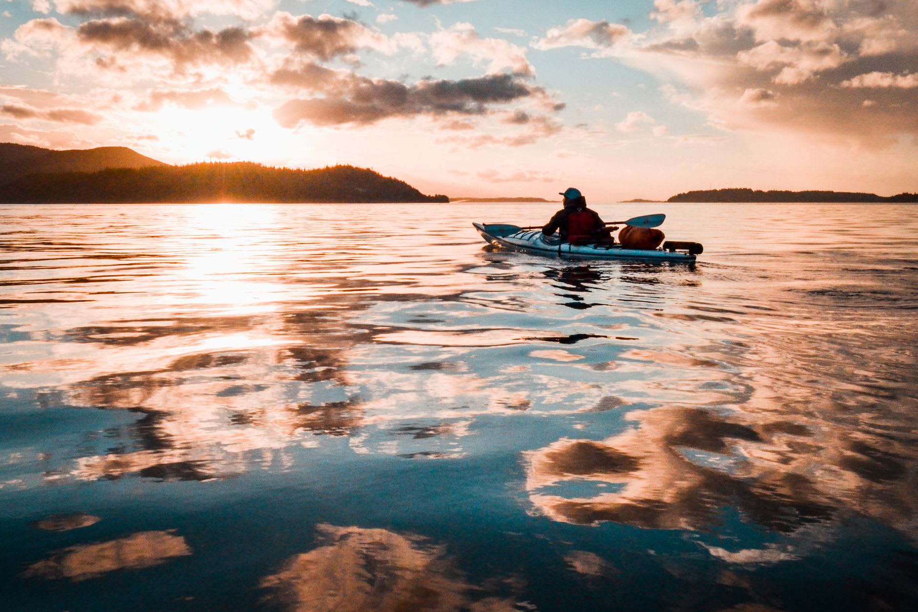 A small wooden sailboat in the calm waters off Salt Spring Island at sunset, the Gulf Islands and distant Vancouver Island peaks going purple in the light