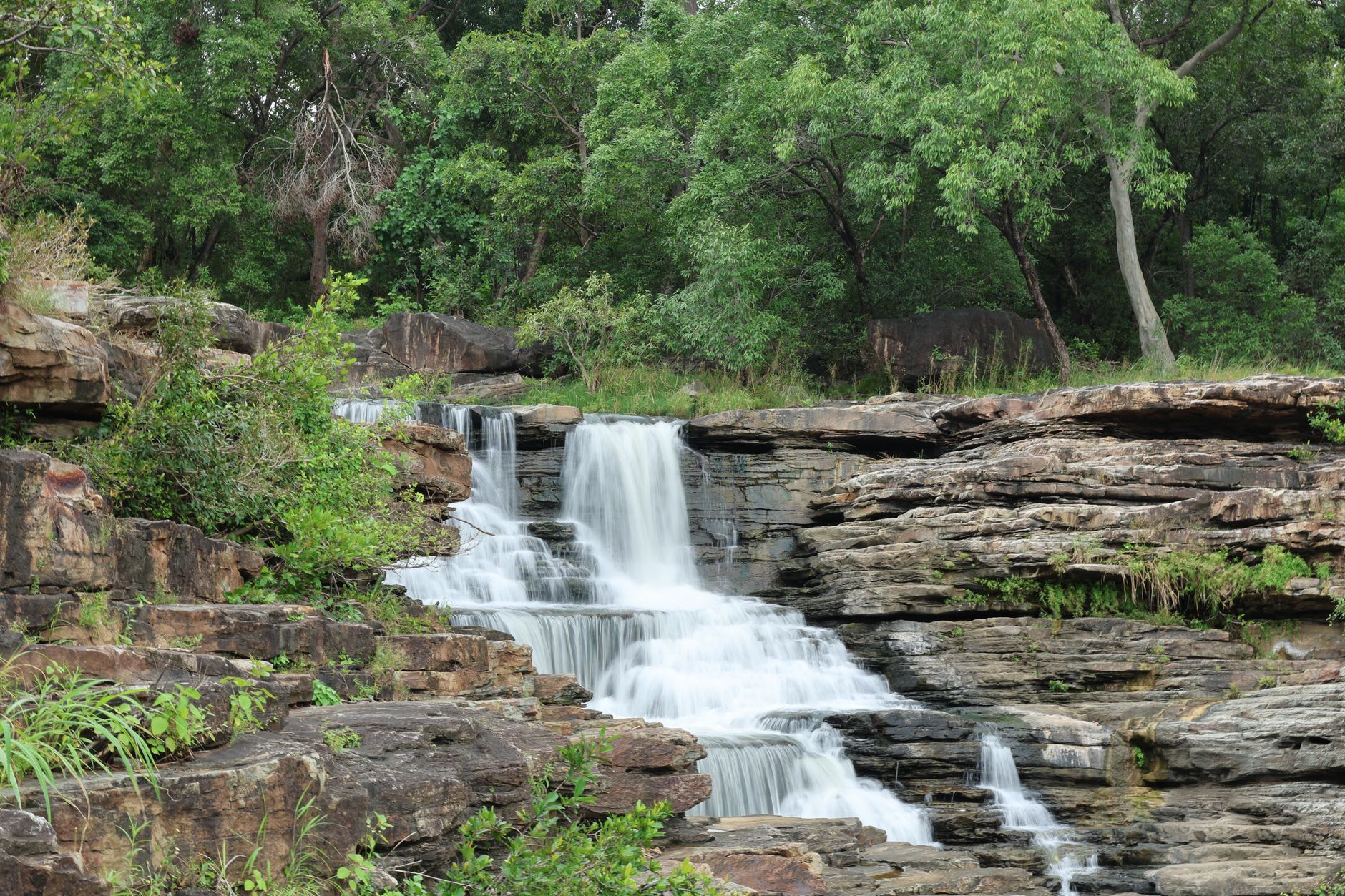 La cascade supérieure de Karfiguéla dévalant un basalte en gradins vers une vasque dans l'ombre, des fougères agrippées à la roche mouillée de chaque côté