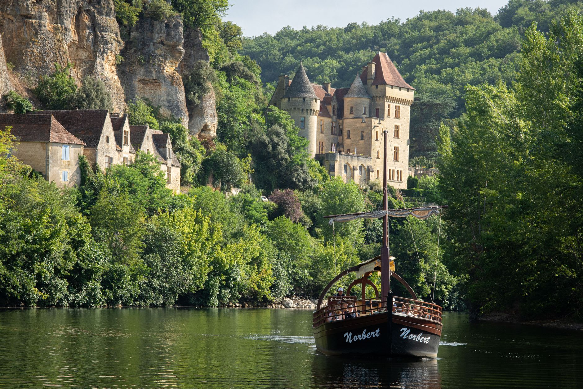 Wooden gabare boats moored along the Dordogne riverbank at La Roque-Gageac with the village and cliff behind