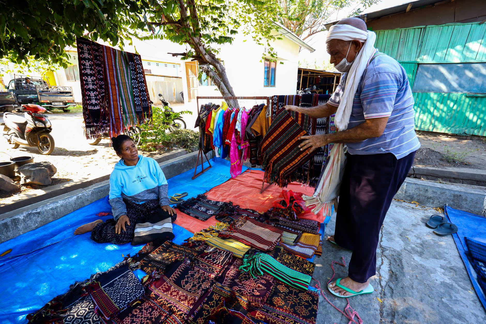 A weaver working a backstrap loom on a village porch near Maumere, an indigo-dyed ikat cloth stretched in front of her