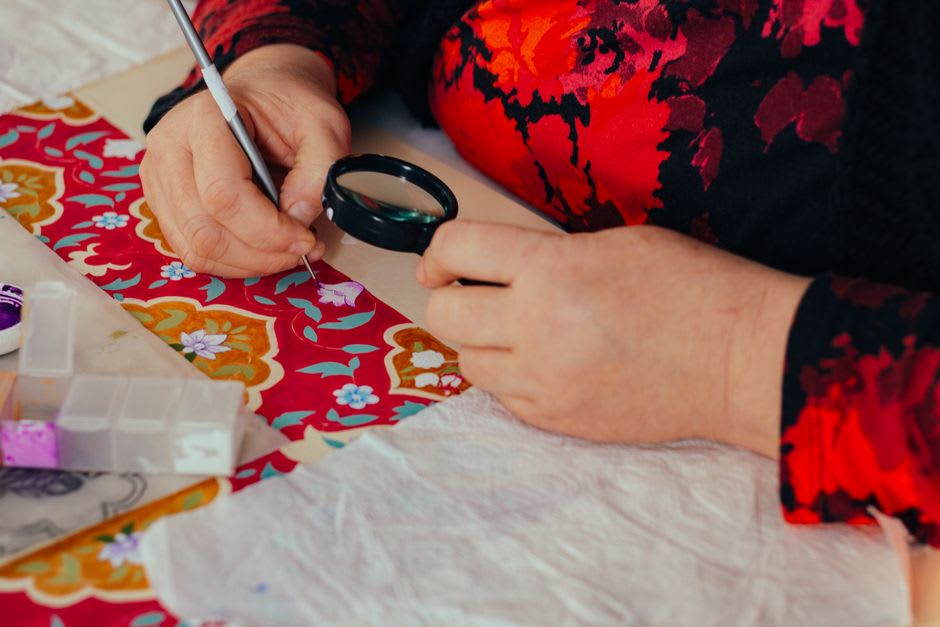 A lacemaker's hands working Alençon needle lace over a pattern under a magnifying lamp