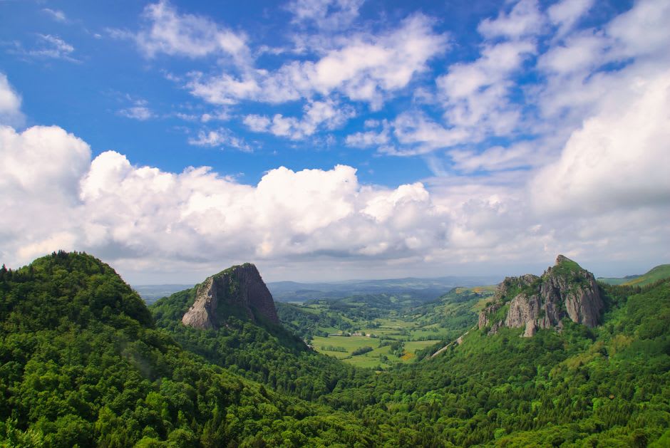 A grassy volcanic hillside trail above Orcival with the basilica's stone towers visible in the valley below