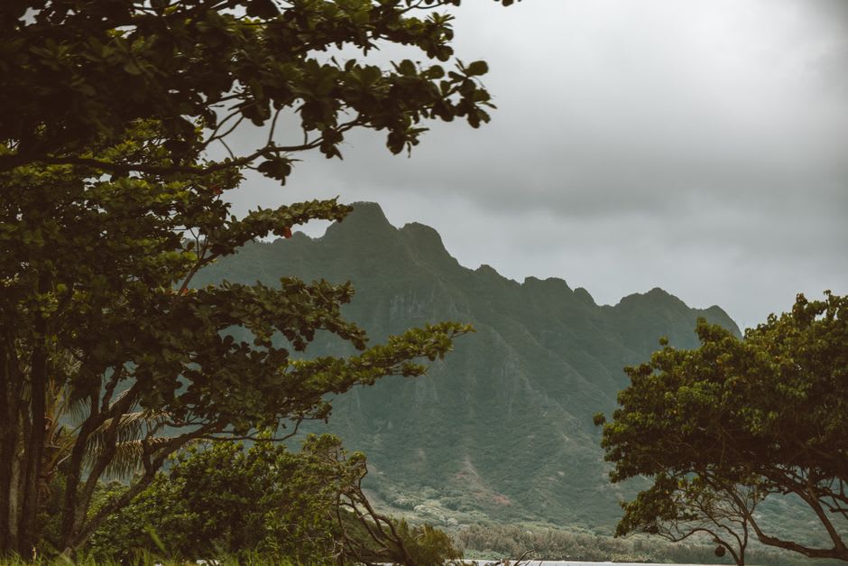 A steep dirt hiking trail climbing through green pastureland with clouds gathering over the West Maui Mountains