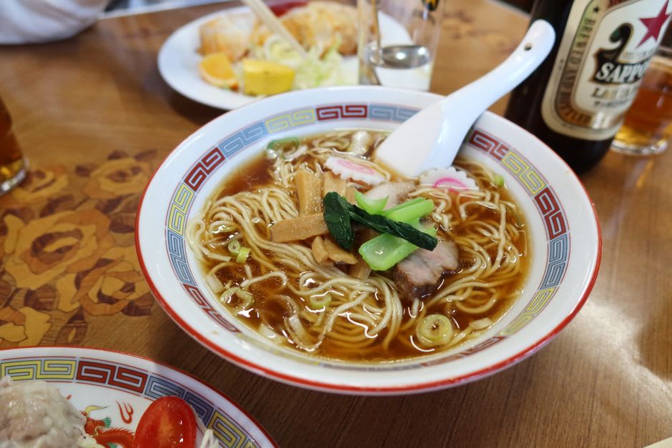 Un tazón de ramen shoyu estilo Asahikawa con fideos delgados y rizados, una capa de aceite sobre el caldo oscuro, y cerdo en rodajas encima