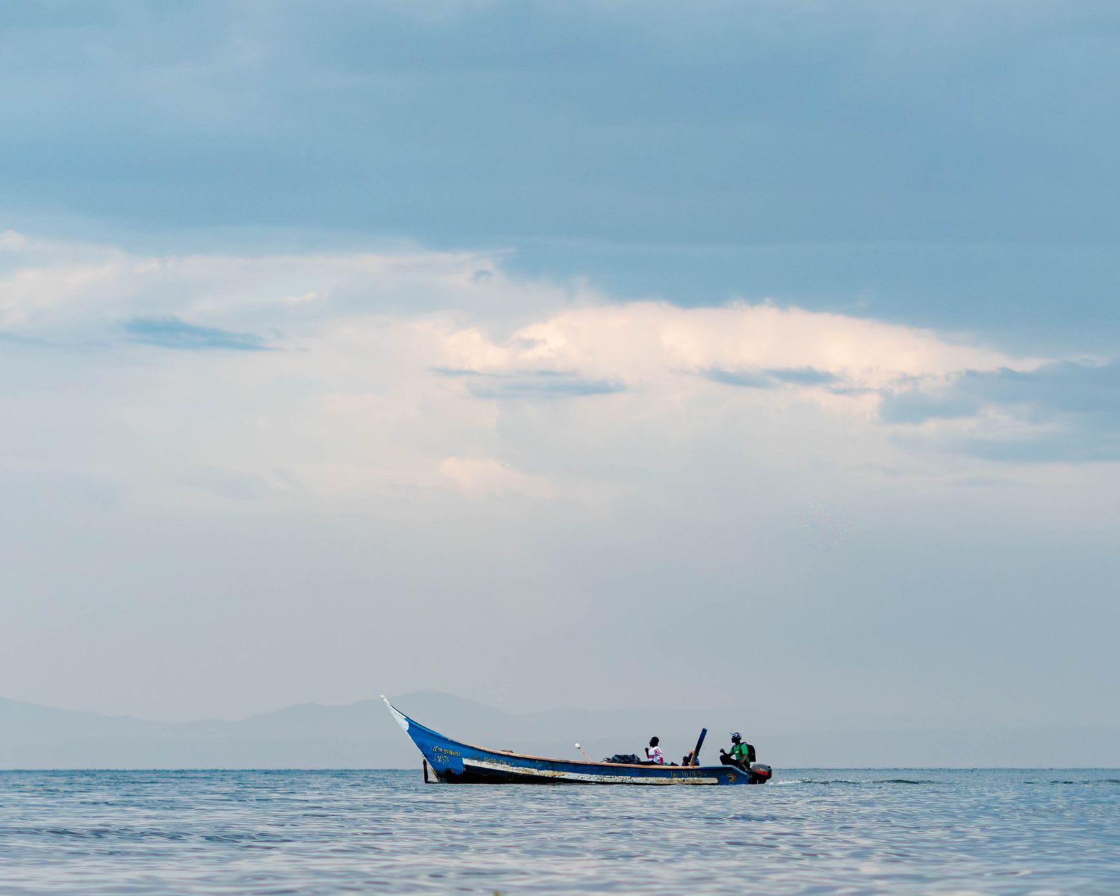 Ekiru, a Turkana elder guide, standing on the black volcanic shoreline of Lake Turkana with his walking stick, looking out across the jade water in the afternoon light