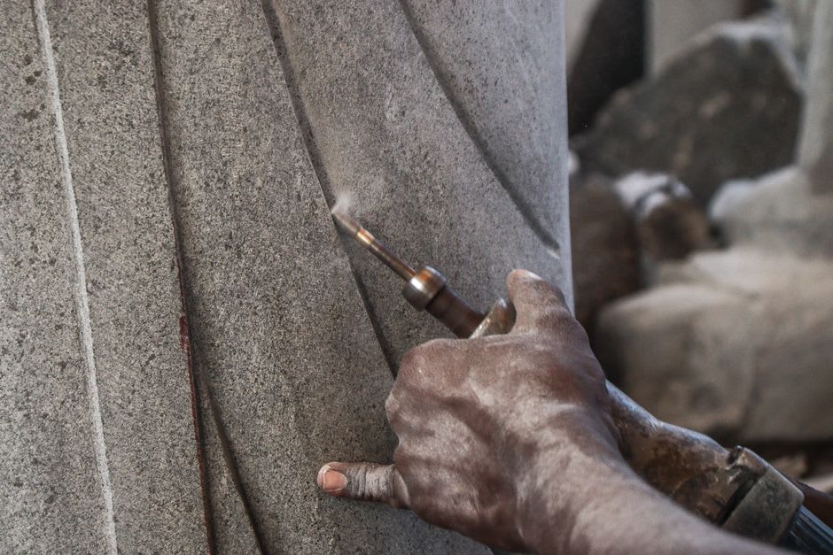 A stone sculptor's hands chiseling a granite deity in a Mahabalipuram workshop, granite dust catching the light