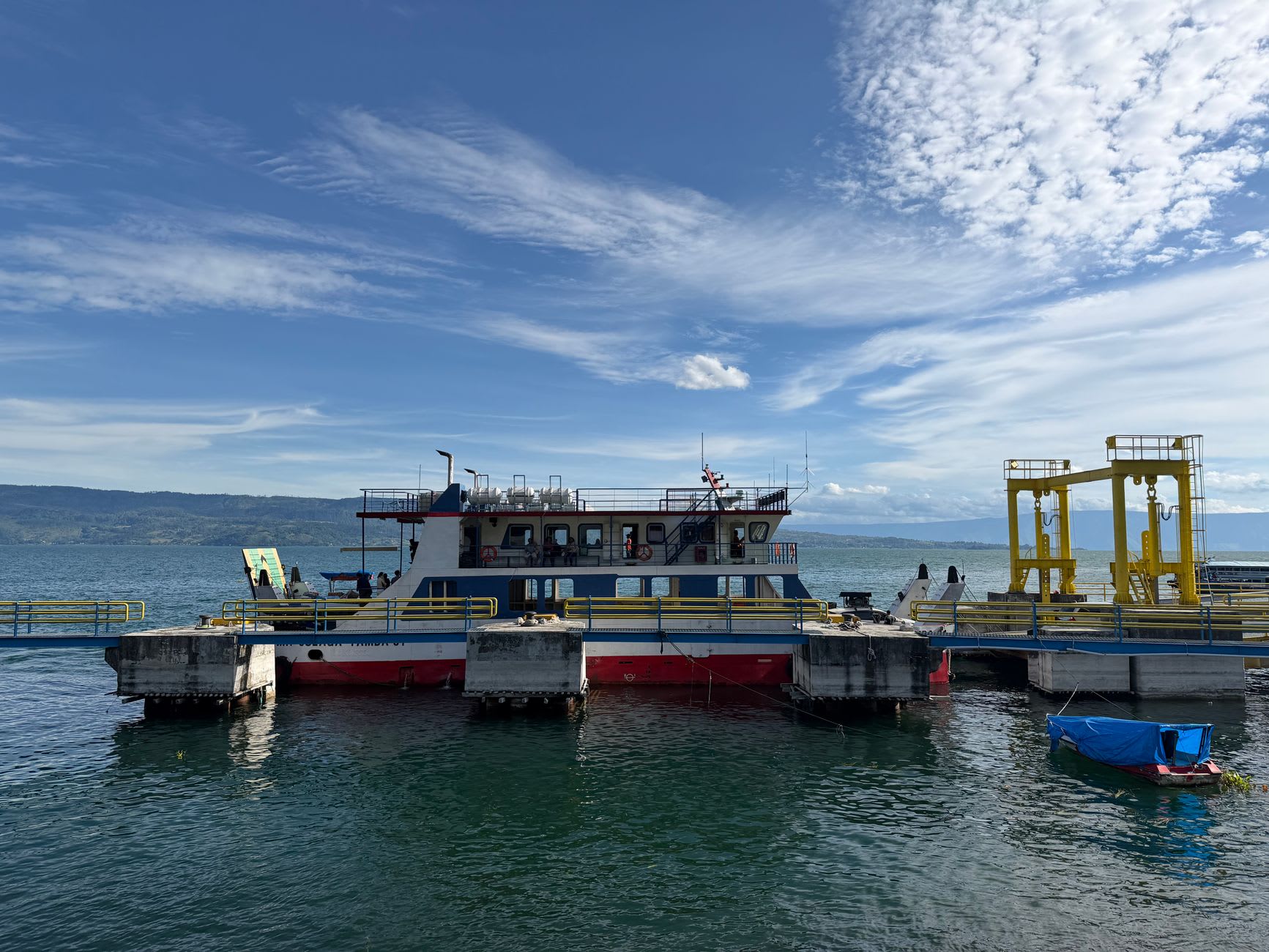 A wooden ferry crossing the calm blue water of Lake Toba toward the green shoreline of Samosir Island, volcanic ridges behind