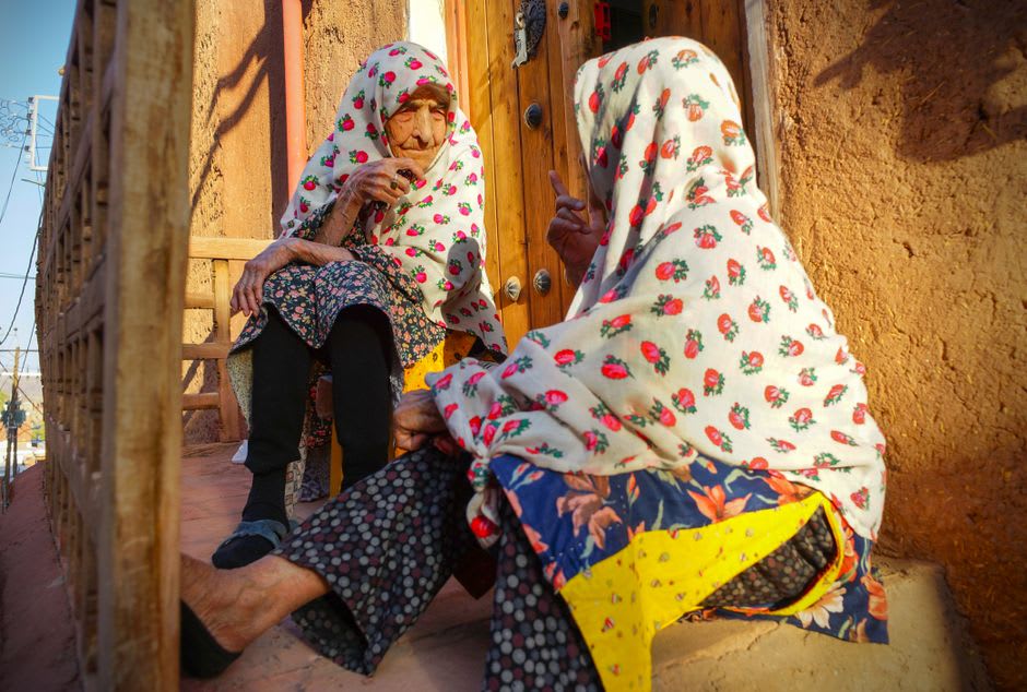 Mujer mayor con pañuelo floreado tradicional y falda plisada de pie en un callejón de arcilla roja en Abyaneh