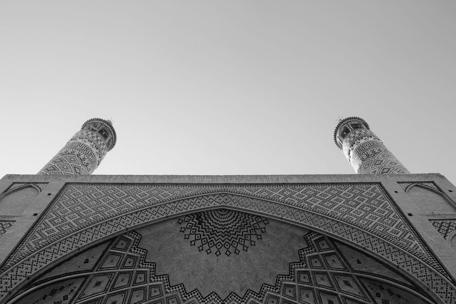 One of the two blue-tiled minarets of Monar Jonban mid-sway, captured with visible motion blur against the sky
