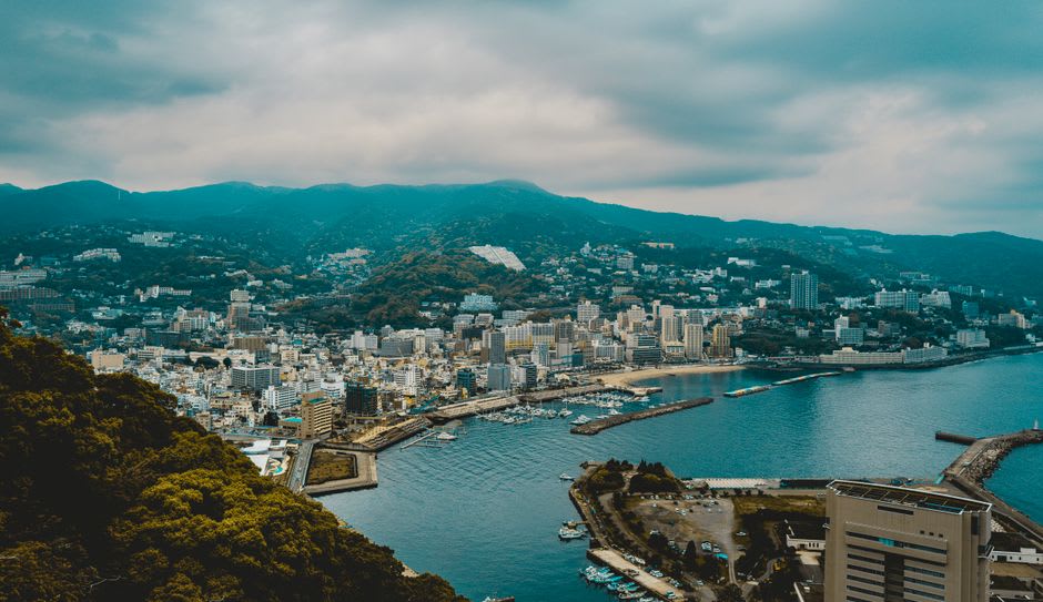 An open-air onsen bath at a hillside Atami ryokan at dusk, steam rising over the water with the curved bay and town lights spread out below