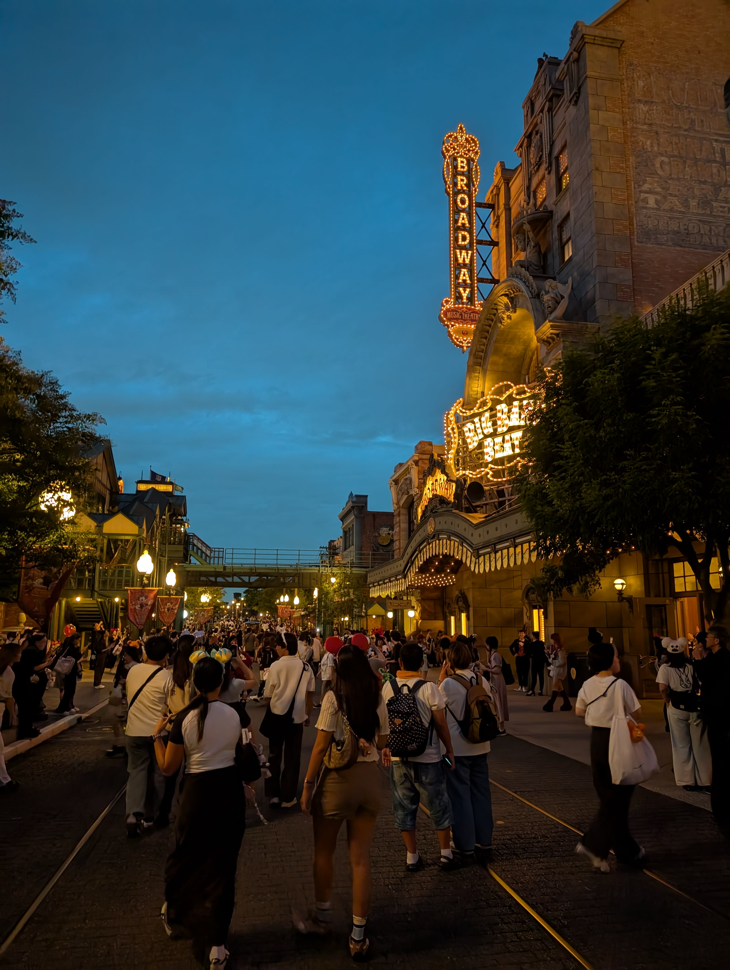 The Broadway district at night — illuminated marquees, golden neon, the Tower of Terror in the distance, crowds walking down a street that belongs to a dream of New York