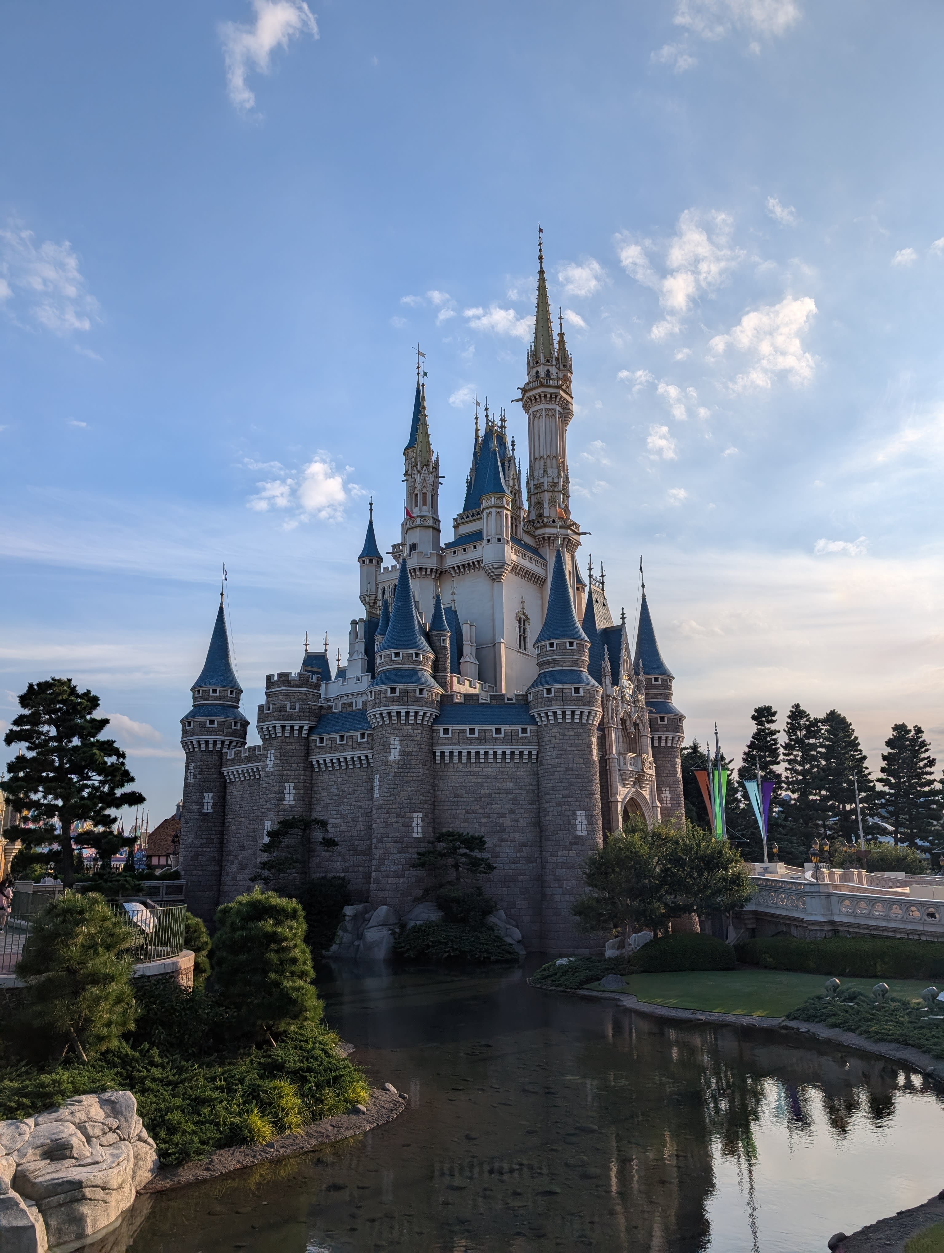 Cinderella Castle at golden hour — seen from behind, across the moat, with Japanese pines framing the spires against a warm sky