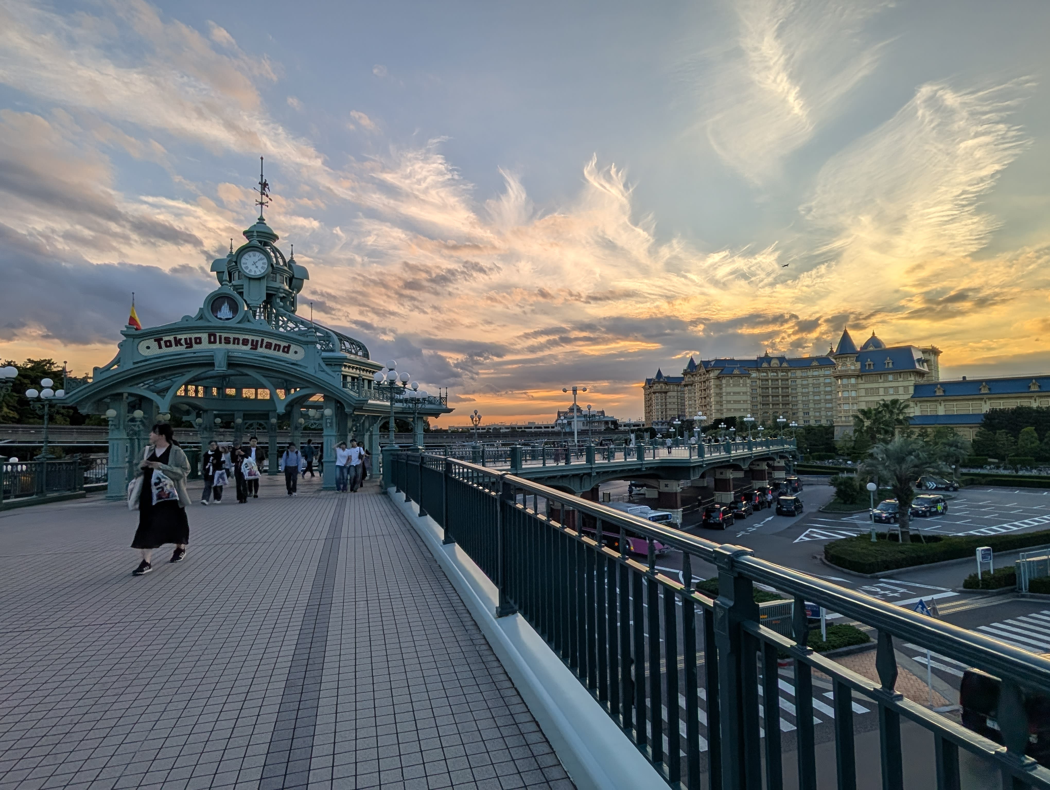 The Tokyo Disneyland entrance arch at sunset — the clock tower silhouetted against an amber sky, the Disney hotel visible beyond, visitors walking home