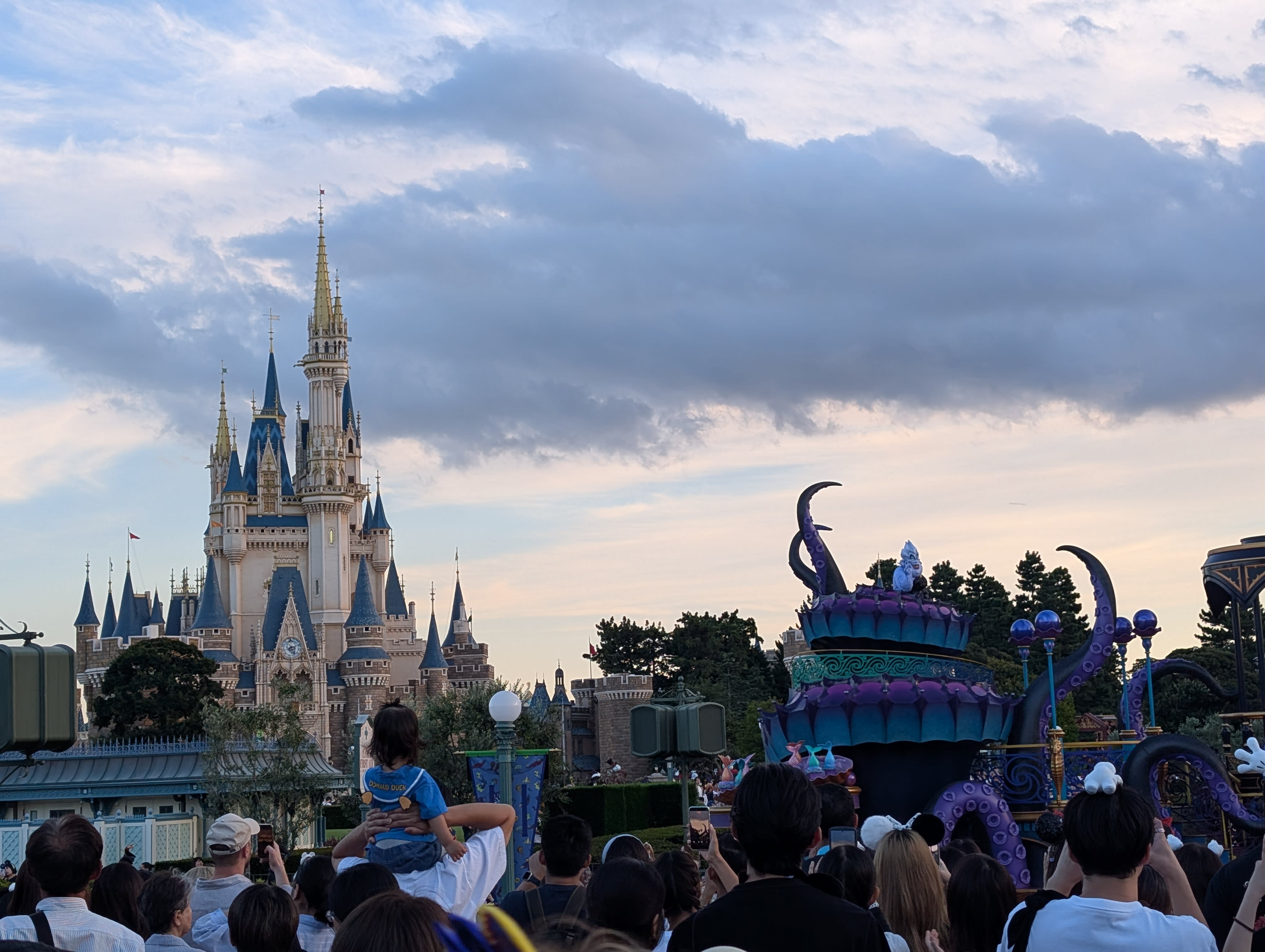 The Halloween parade at dusk — Ursula's purple tentacle float passing before Cinderella Castle, the crowd watching, a child on his father's shoulders