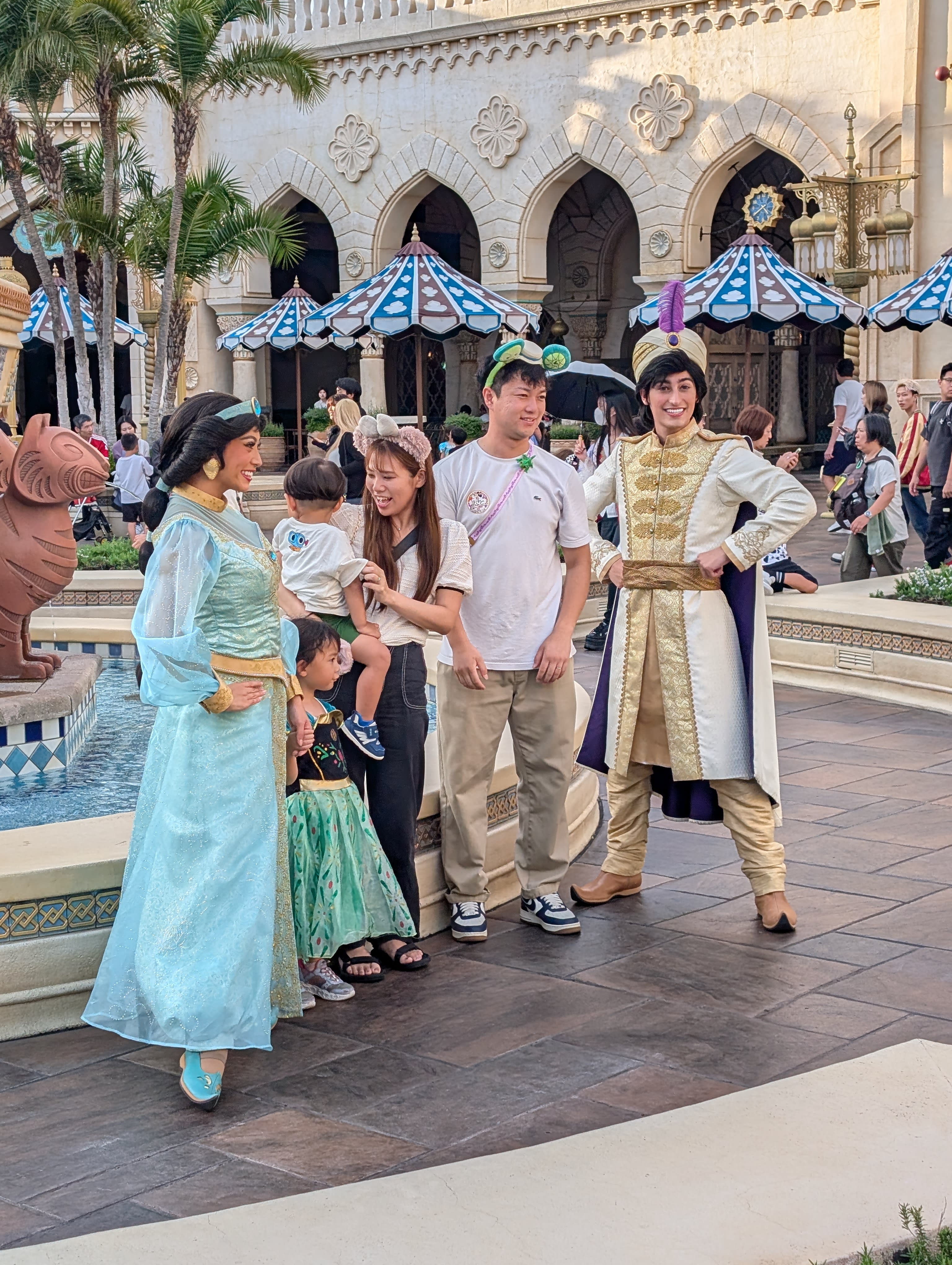 Jasmine and Aladdin with a Japanese family at the Arabian Coast — the little girl dressed as Jasmine, Aladdin kneeling, the palace arches behind them