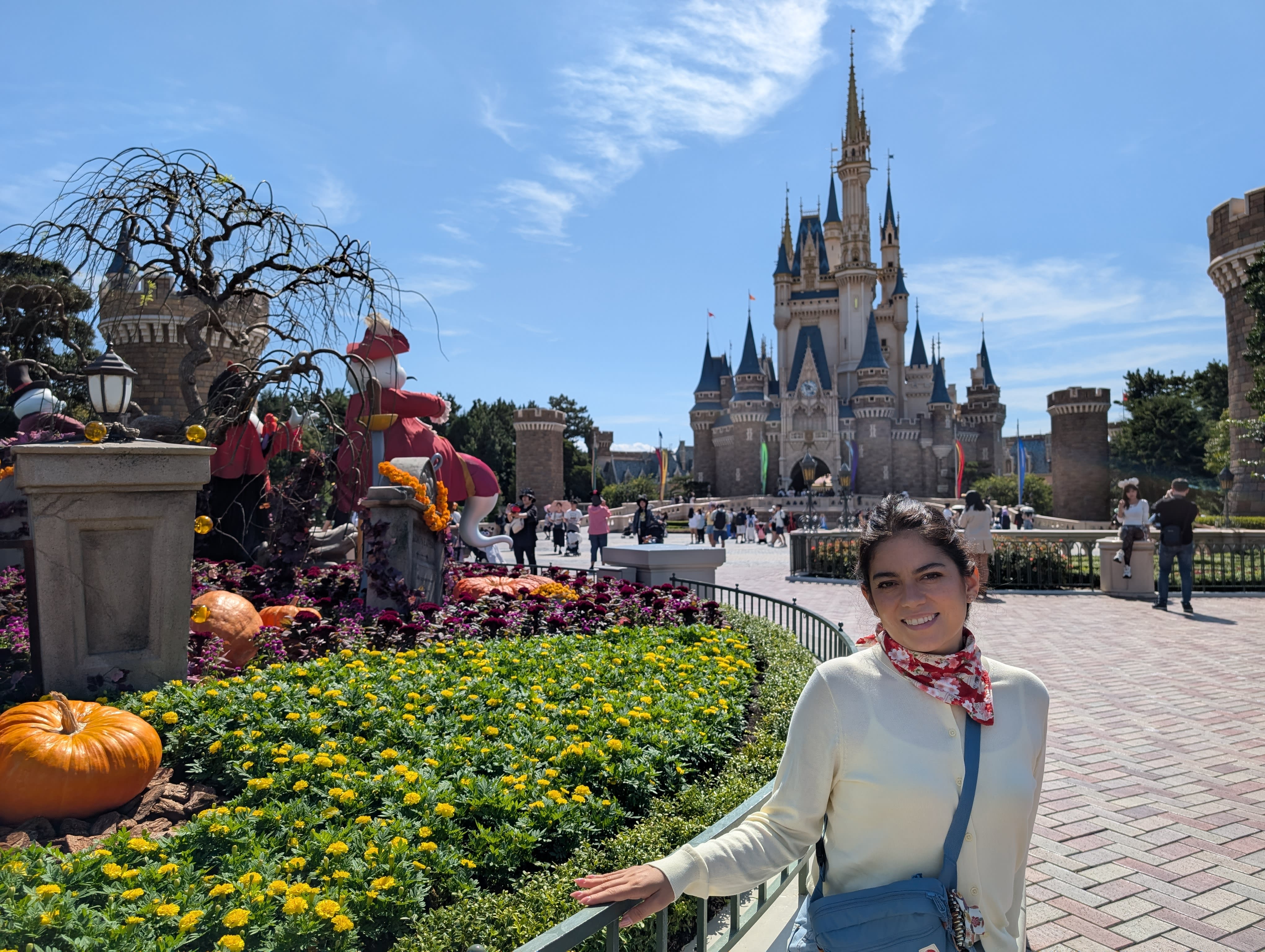 Lia in front of Cinderella Castle at Tokyo Disneyland — Halloween pumpkins and flowers in the foreground, the castle soaring behind her against a blue sky