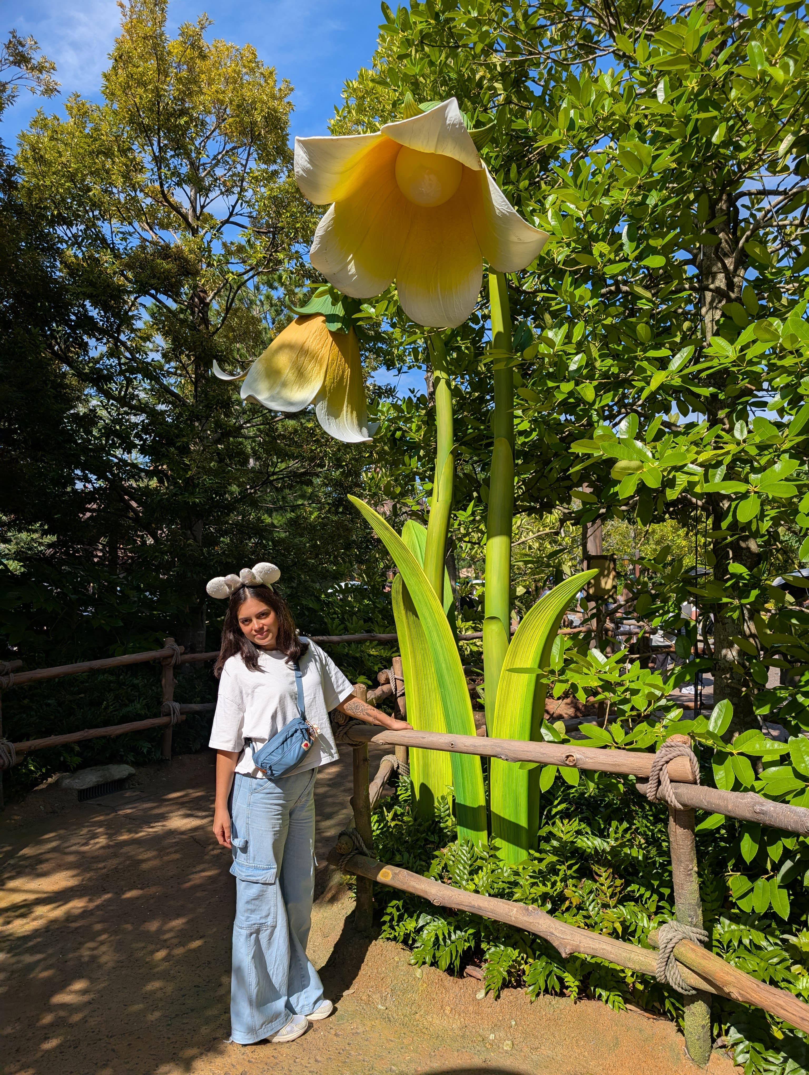 Lia standing next to a giant daffodil in Fantasy Springs — the flower towering above her, sunlight filtering through the petals, the forest stretching behind