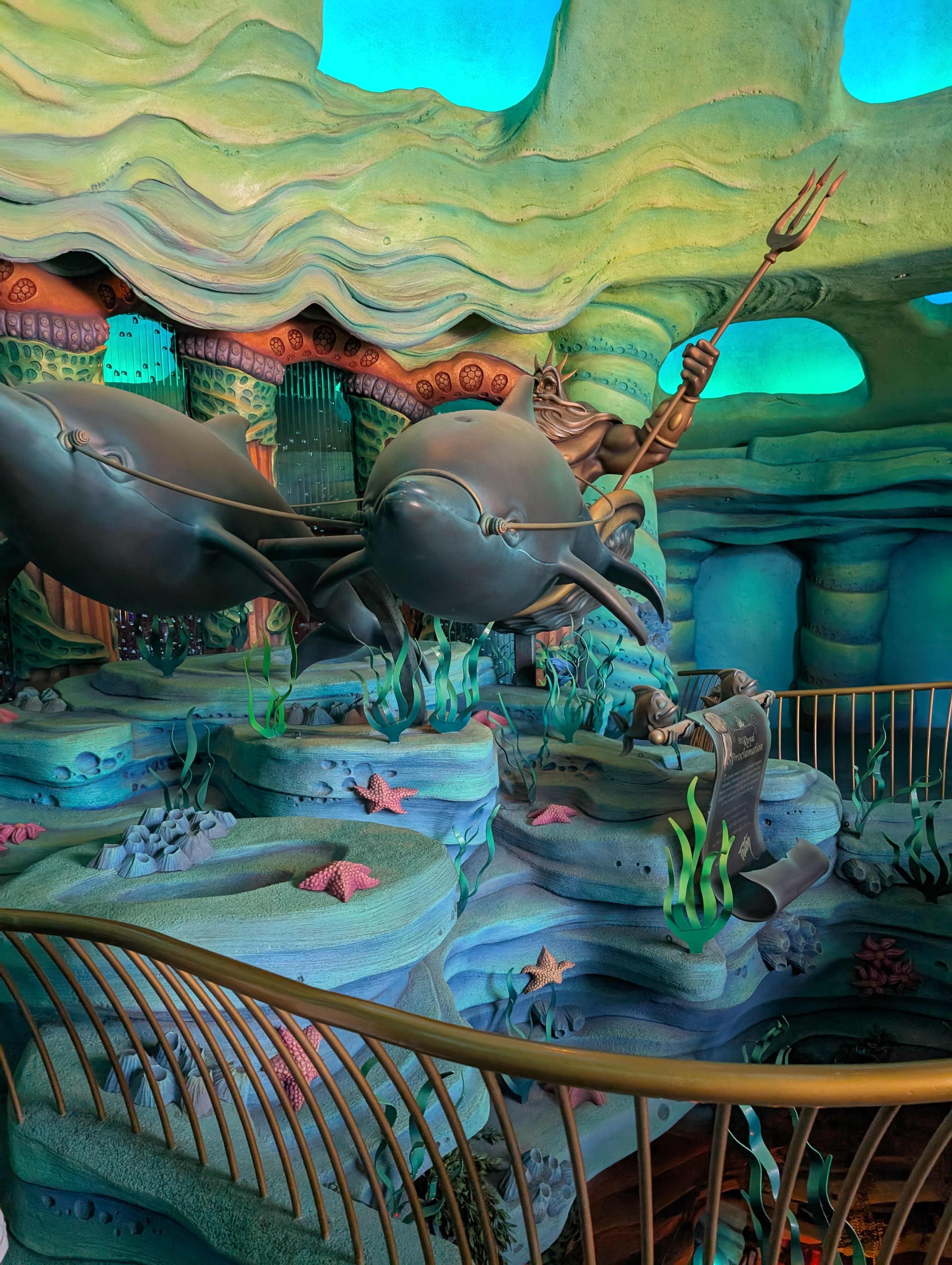 King Triton's sculpture in Mermaid Lagoon — trident raised, dolphins leaping, surrounded by coral and underwater scenery in blue and green light