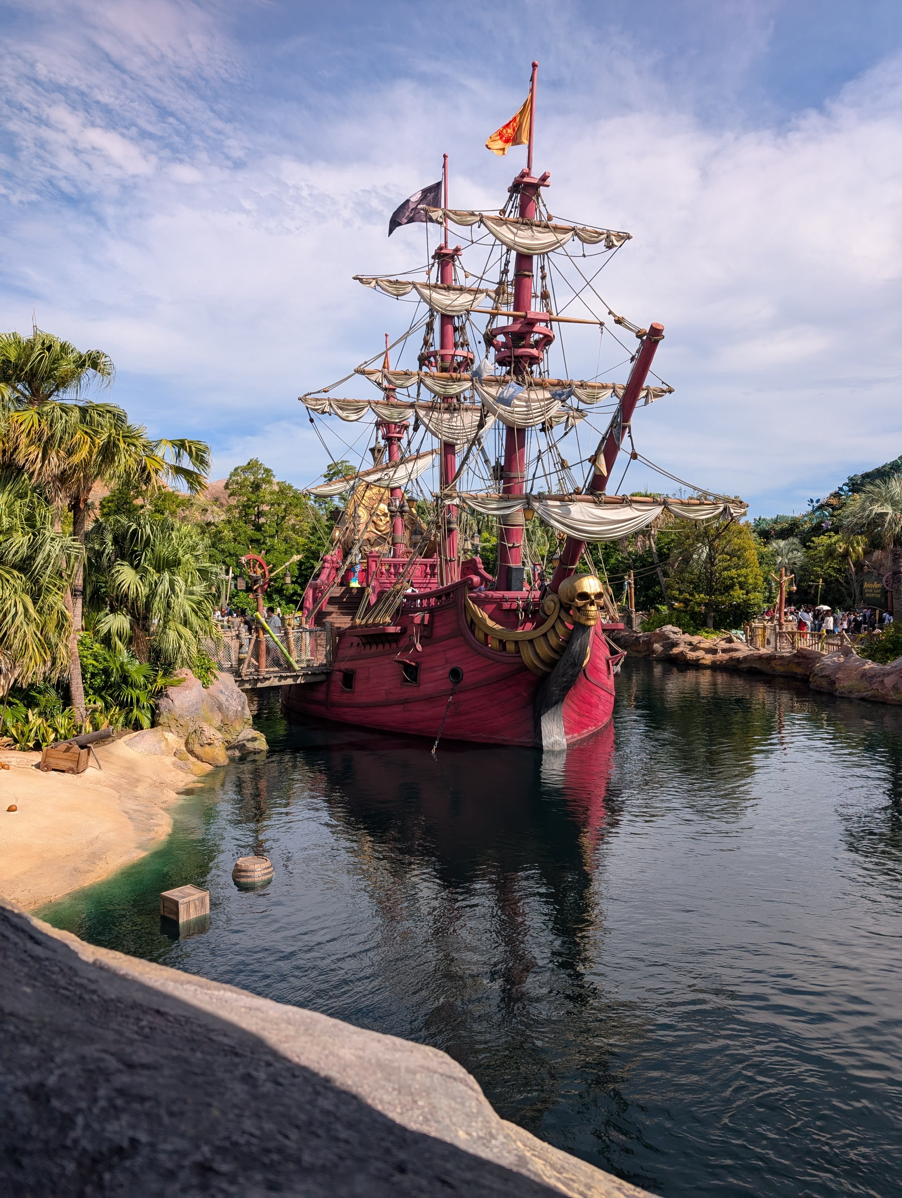 Captain Hook's pirate galleon moored in the harbour — red hull, full rigging, skull figurehead, palm trees and clear water, a prop that has forgotten it is a prop