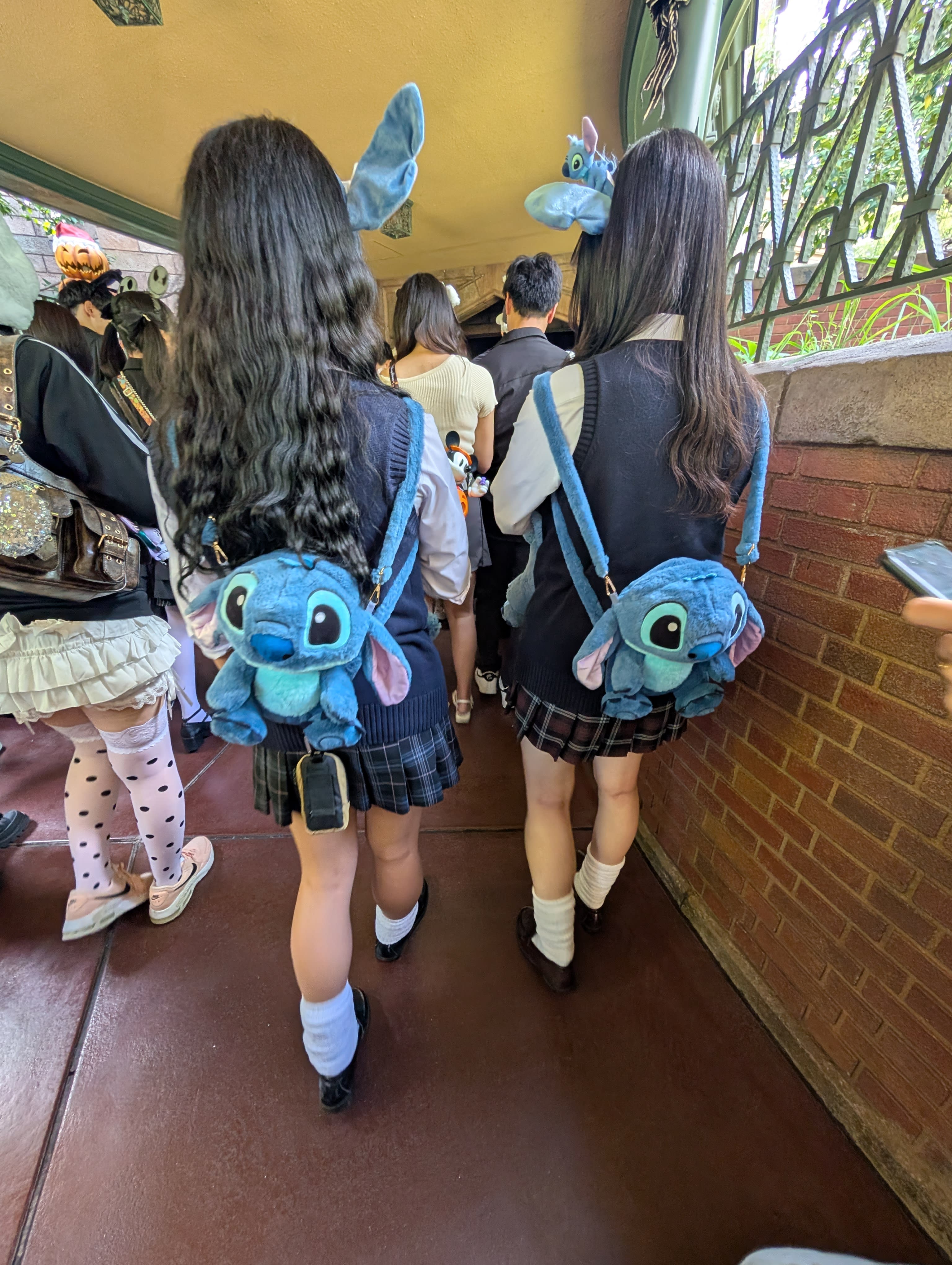 Two Japanese girls with matching Stitch ear headbands and Stitch plush backpacks walking through the park — coordinated down to the last detail