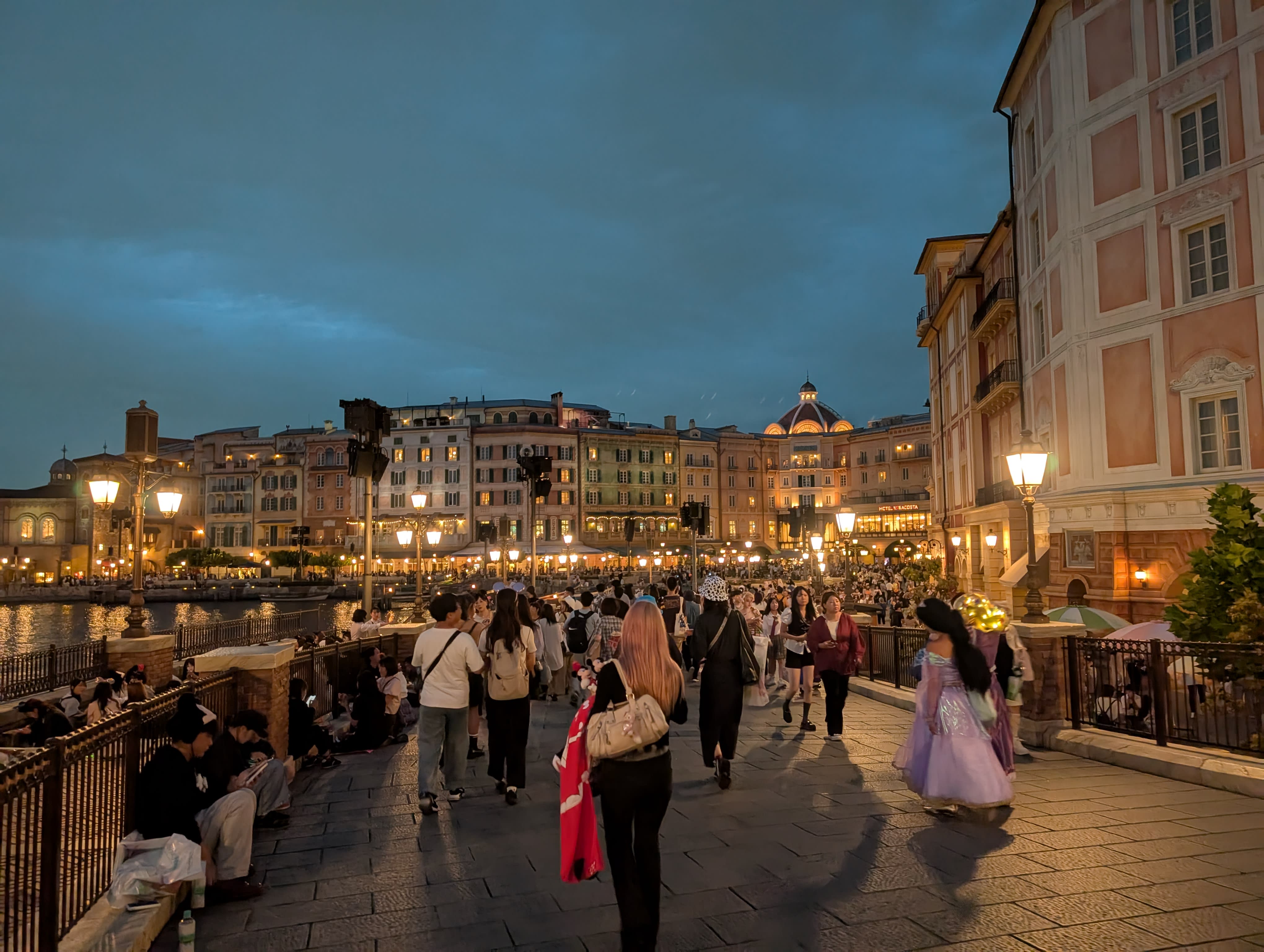 The Venetian waterfront at DisneySea at dusk — pastel buildings, lamp-lit promenades, visitors in costume walking along the harbour, the sky deep blue