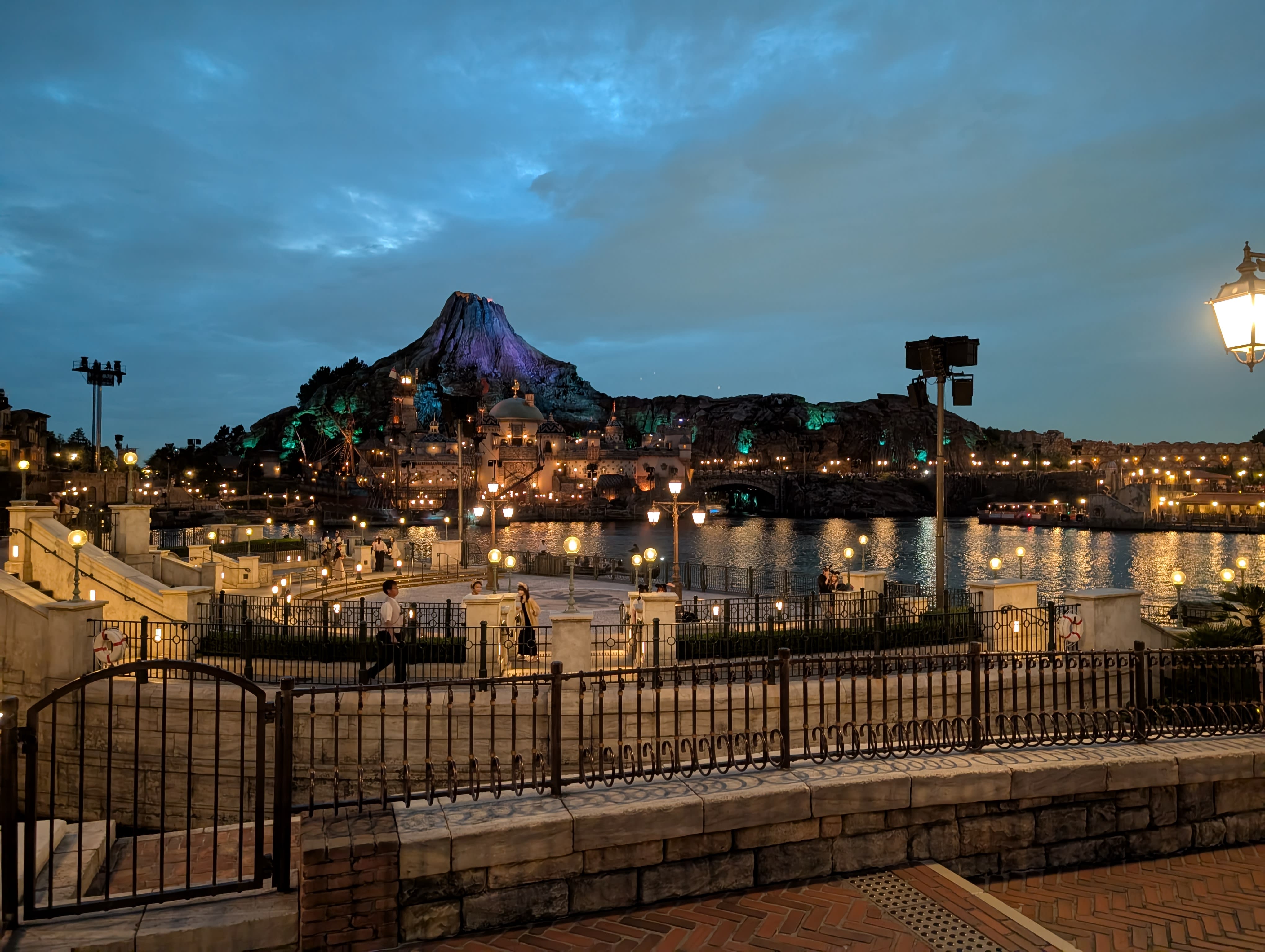 Mount Prometheus and the Mediterranean Harbor at night — the volcano glowing against the sky, stone bridges and lampposts reflected in the dark water