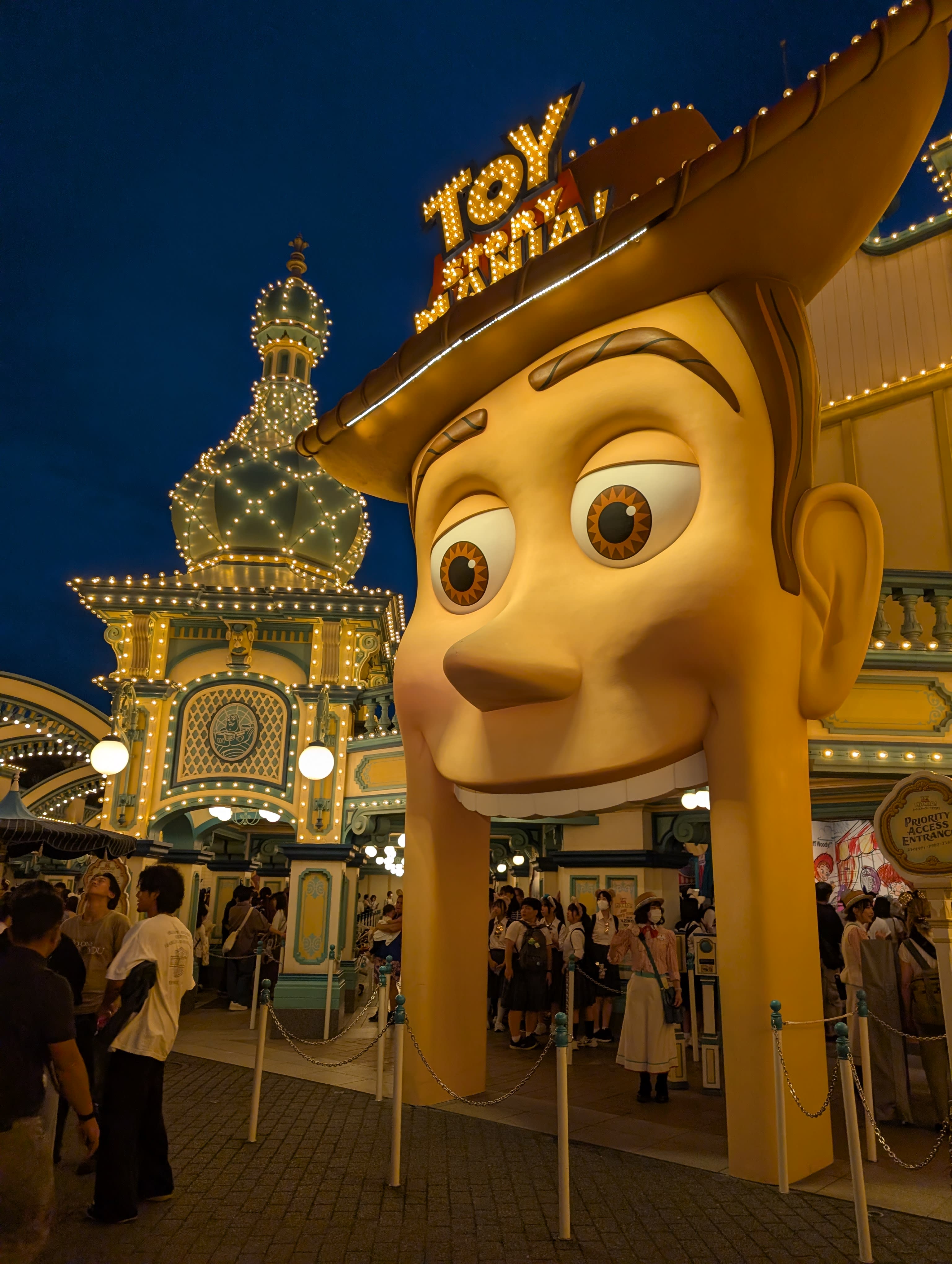 Giant Woody entrance to Toy Story Mania at night — carnival lights, the crowd streaming past, the atmosphere of a world designed to make you feel ten years old