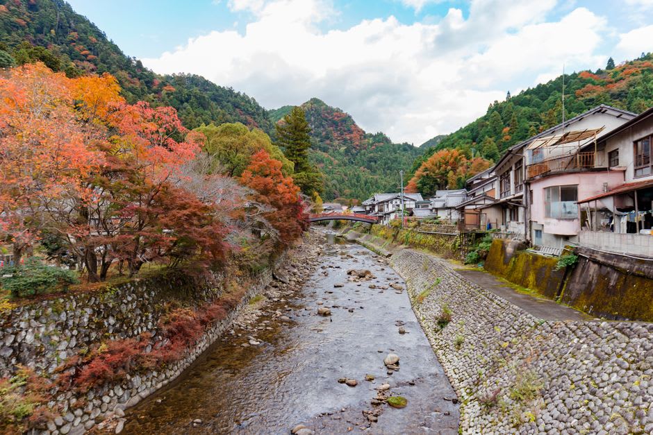 A row of people soaking their feet at a free riverside foot bath in Gero, mountains and a stone bridge behind them