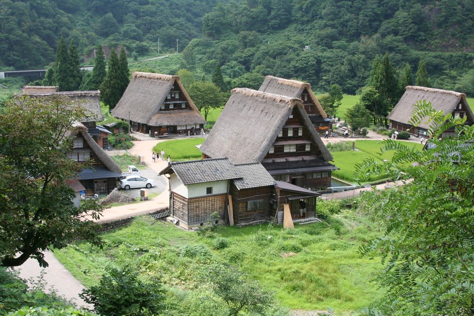A gasshō-zukuri farmhouse in Gokayama under deep winter snow at dusk, warm light glowing from its windows and the steep thatched roof heaped white