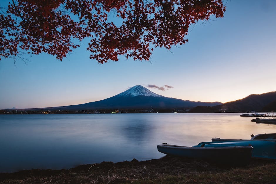 Mt. Fuji rising above Lake Kawaguchiko at twilight, autumn leaves framing the scene in red and gold, the mountain reflected in still water