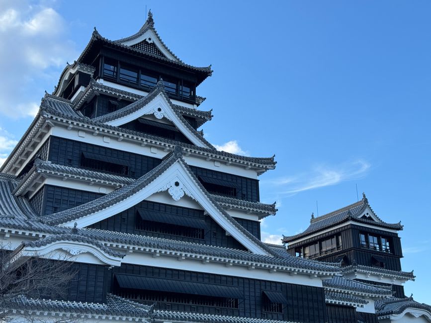 The reconstructed main keep of Kumamoto Castle, black and white against a blue sky, rising above its immense flaring stone walls
