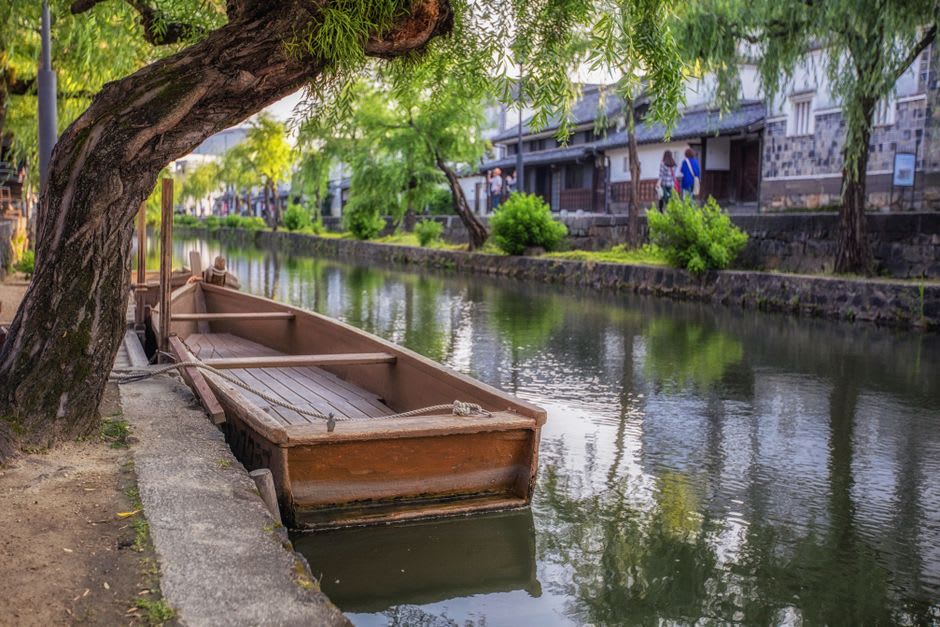 The willow-lined Bikan canal in Kurashiki, white-walled warehouses with black-tiled lower walls reflected in the still green water, a stone bridge arching across