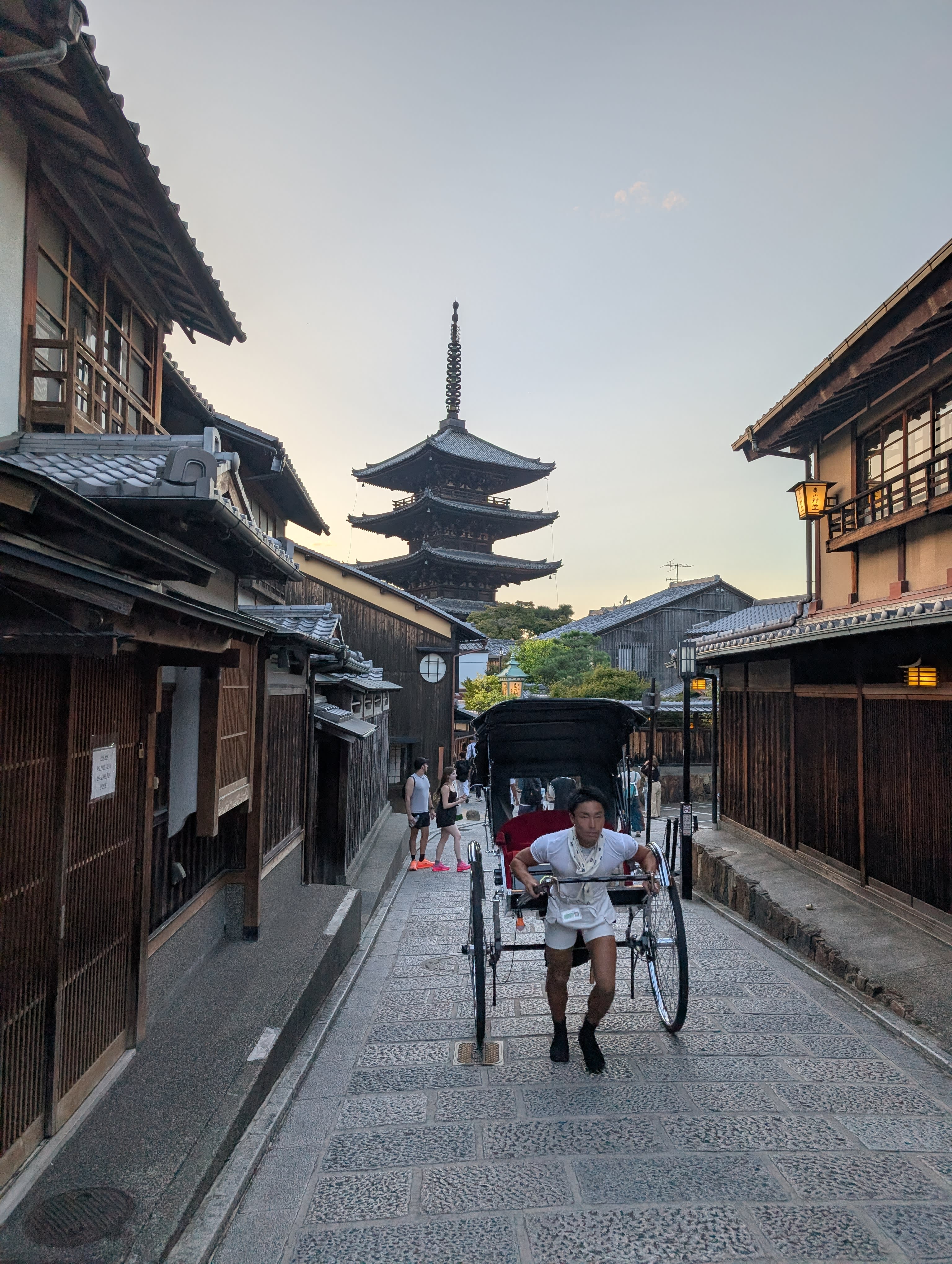 A rickshaw runner pulling passengers through a narrow Higashiyama lane, the five-storey Yasaka Pagoda towering behind him