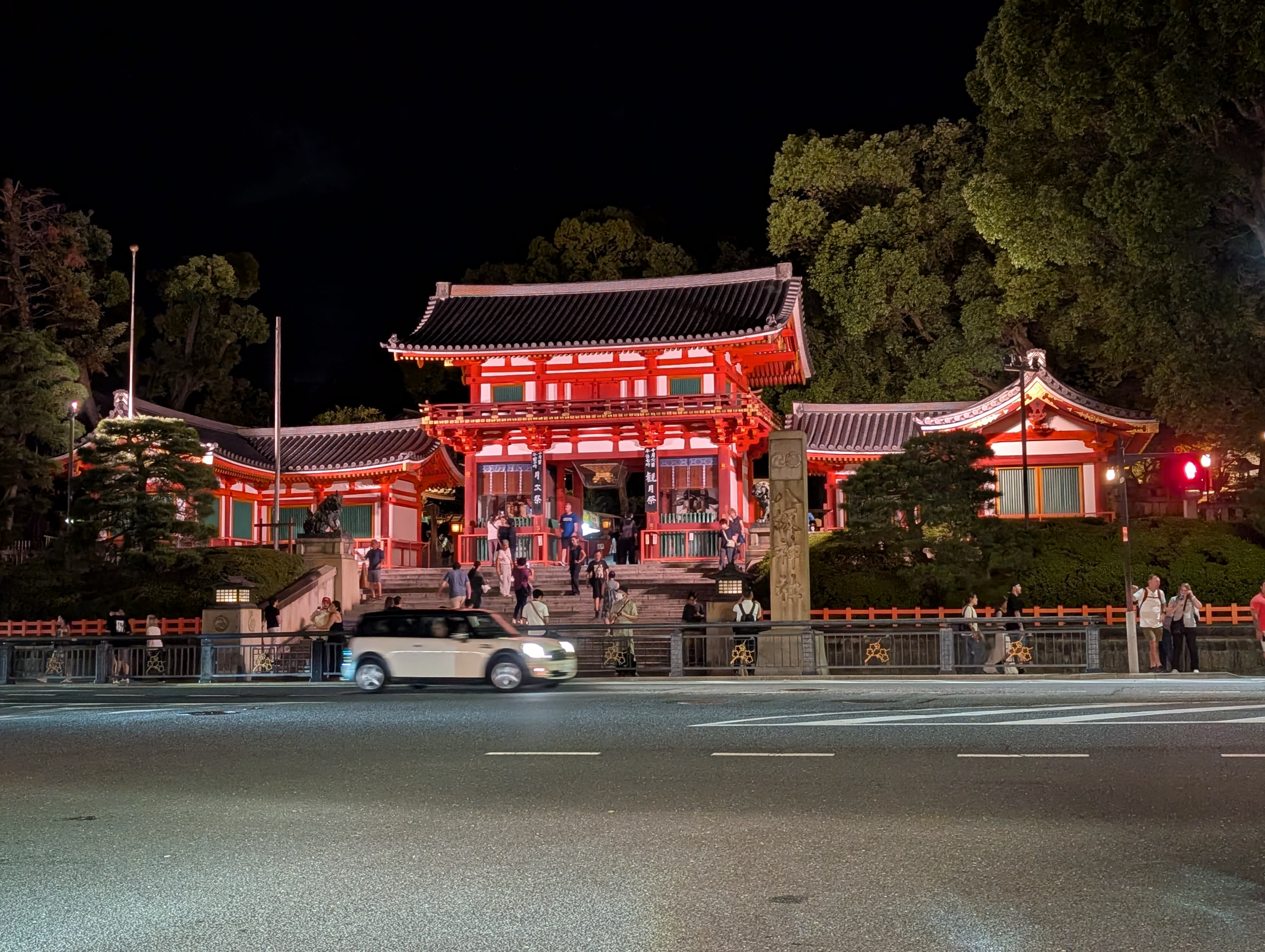 Yasaka Shrine illuminated at night — the vermillion gate glowing against the dark sky, visitors passing through in golden light