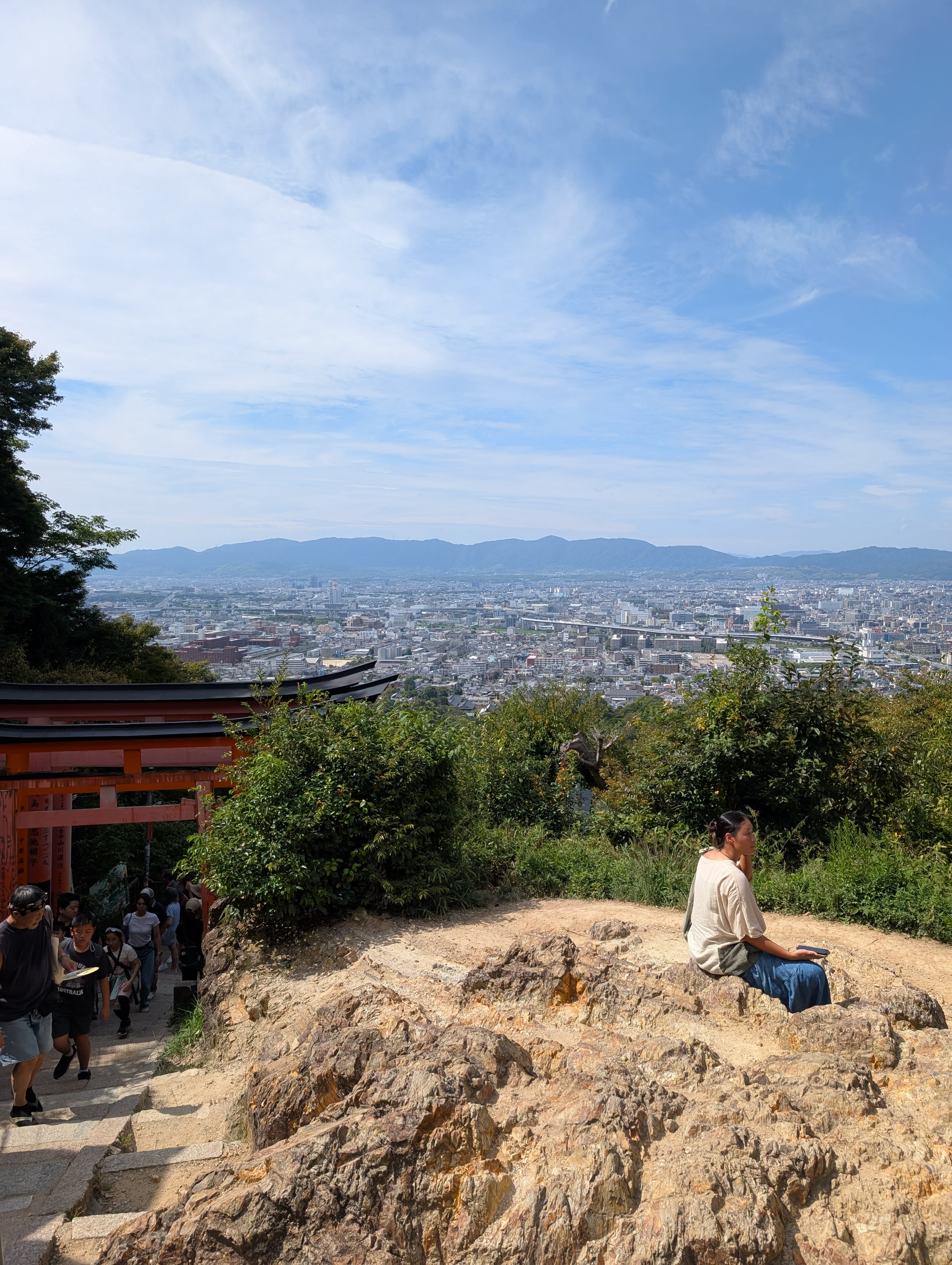 Lia sitting on a rock at the summit of Fushimi Inari, the entire city of Kyoto spread below her, a red torii gate at the edge of the clearing