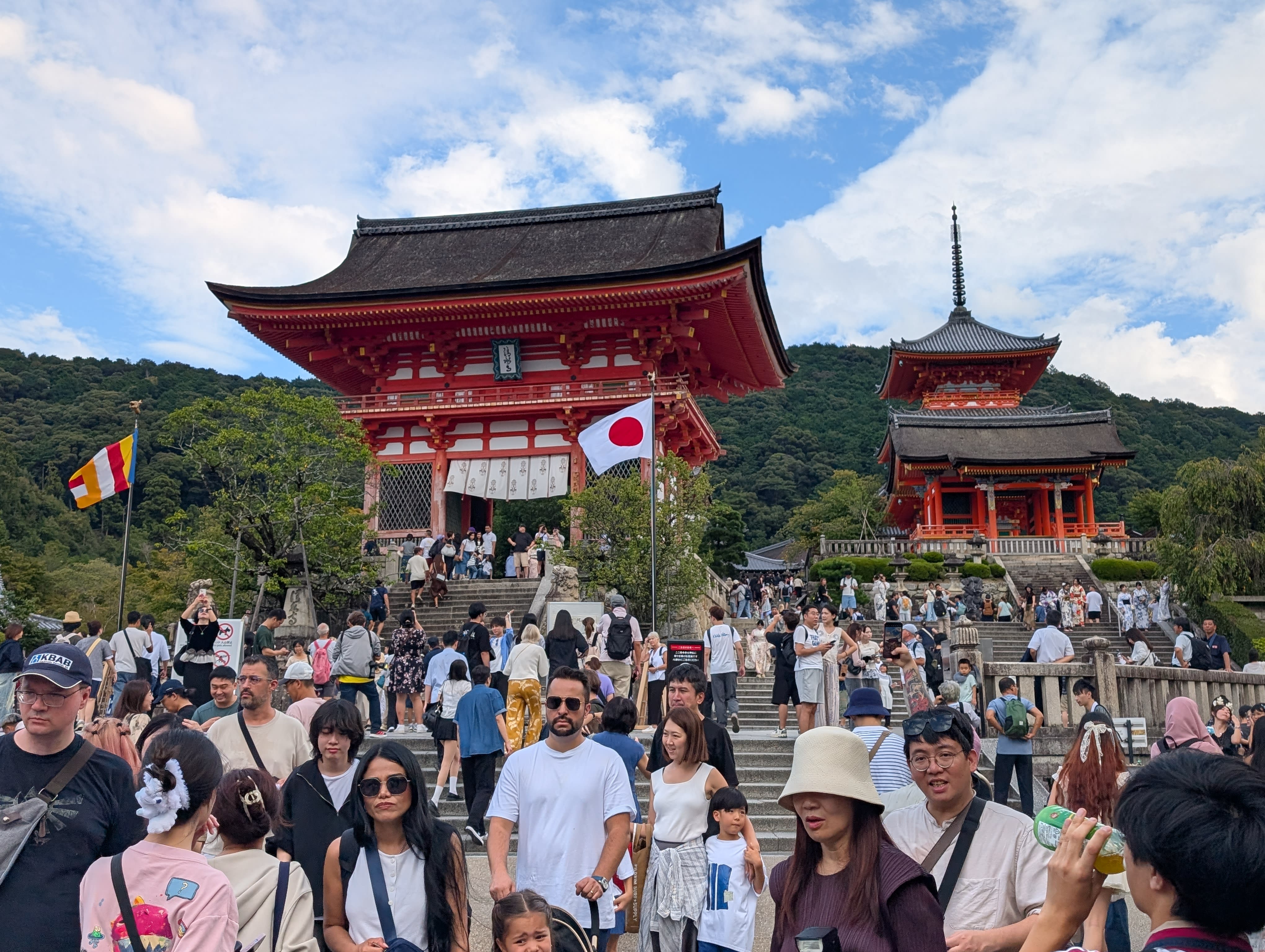 The main gate of Kiyomizu-dera — the massive red-painted Niomon gate with its pagoda, crowds ascending the stone steps, a Japanese flag catching the wind
