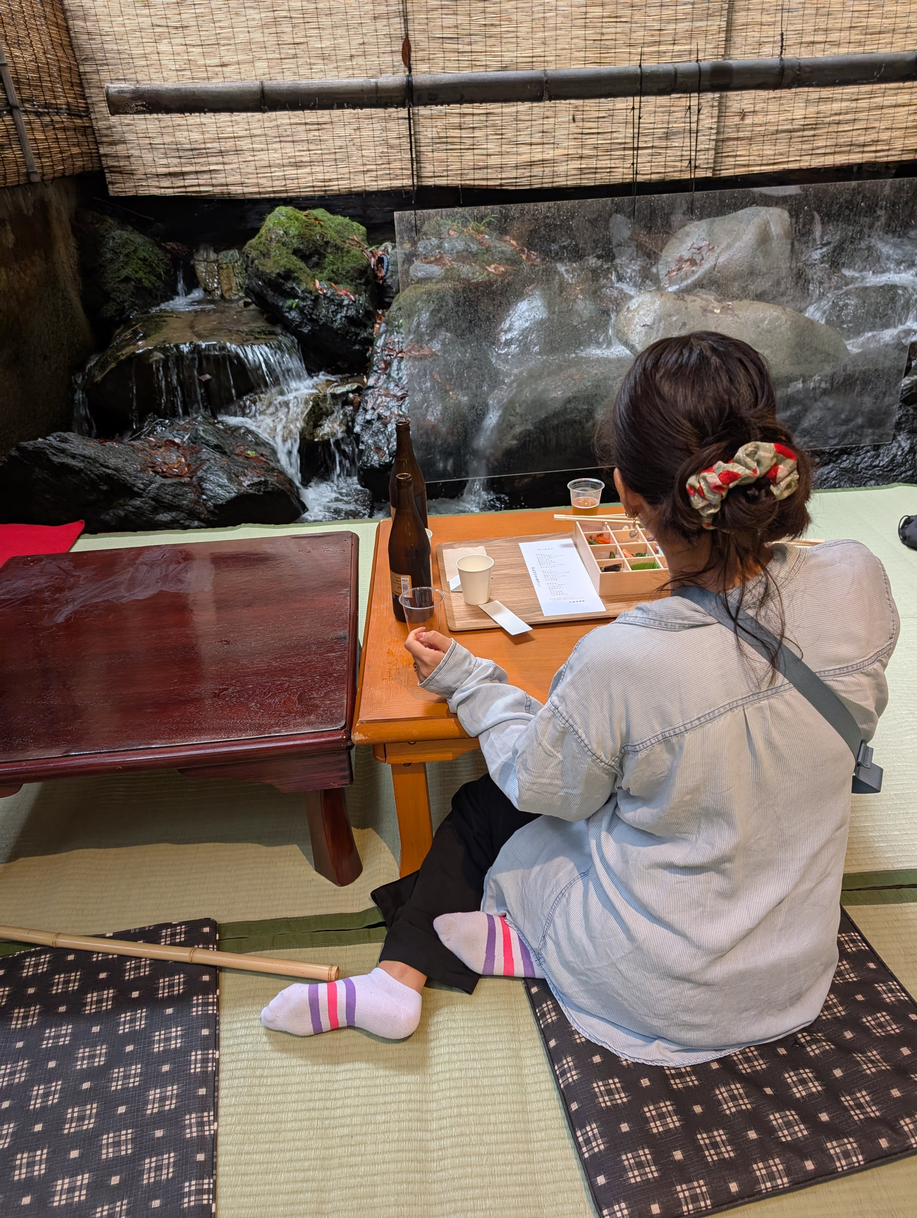 Lia at the kawadoko restaurant in Kibune — seated on tatami at a low table, a bento box and beer before her, a waterfall cascading over rocks just inches away