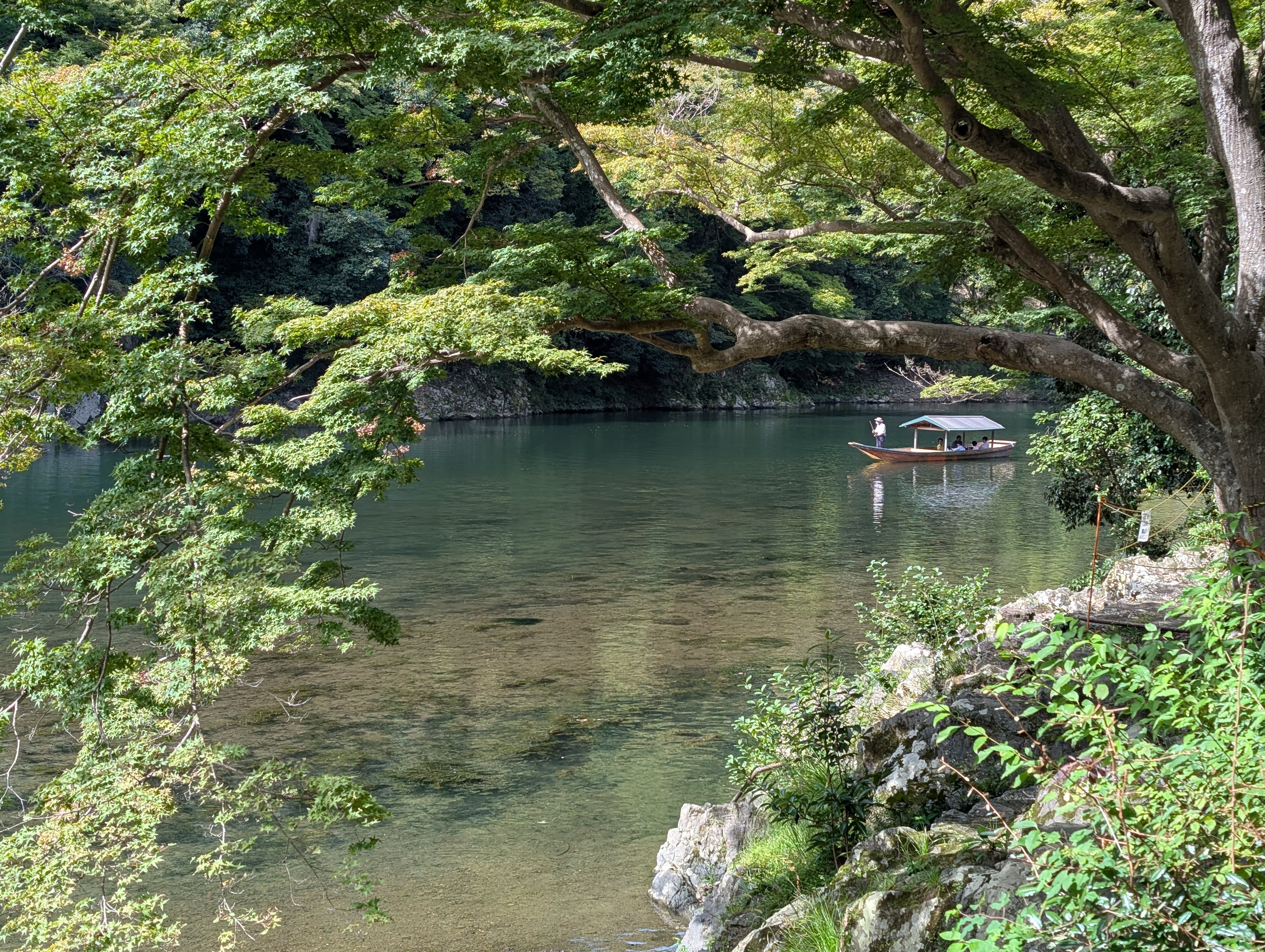 A traditional wooden boat gliding across the pond at Tenryu-ji — the water perfectly still, green trees reflected, the mountains behind