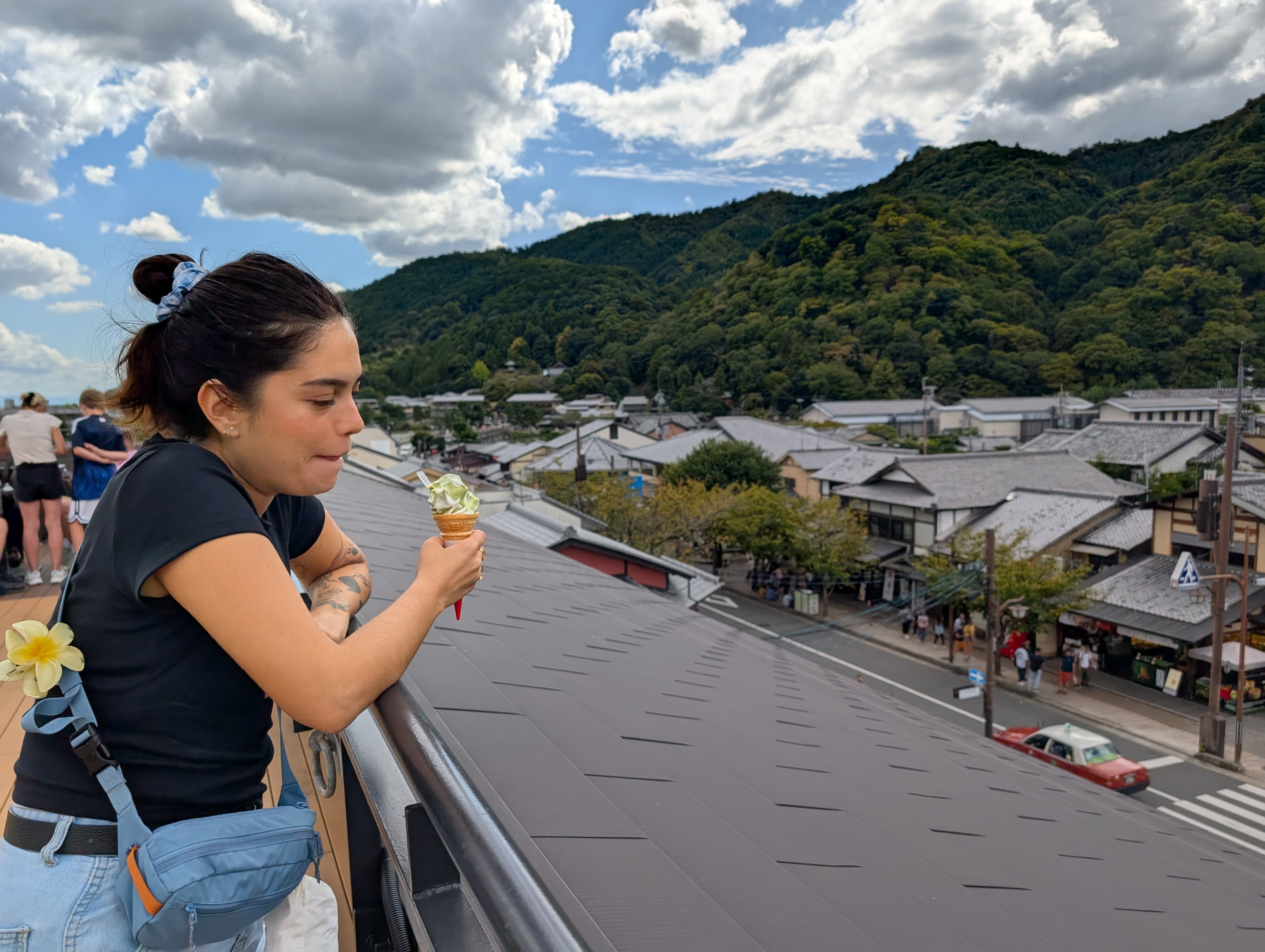 Lia eating matcha ice cream at an Arashiyama viewpoint — leaning on the railing, the traditional rooftops and green mountains of western Kyoto spread below her