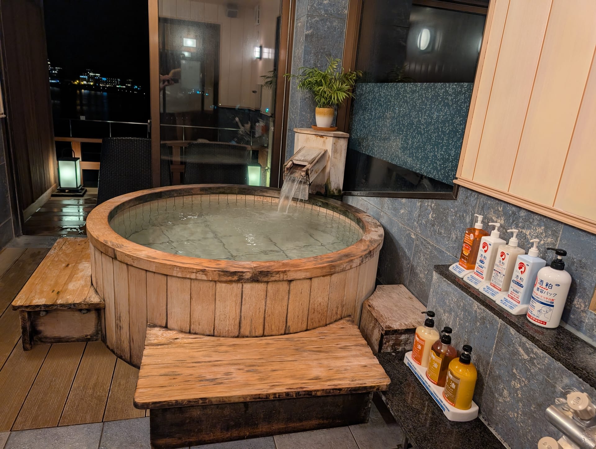 A private wooden onsen bath at Hotel Kasuitei Ohya, water flowing in, with the lake visible through the window at night