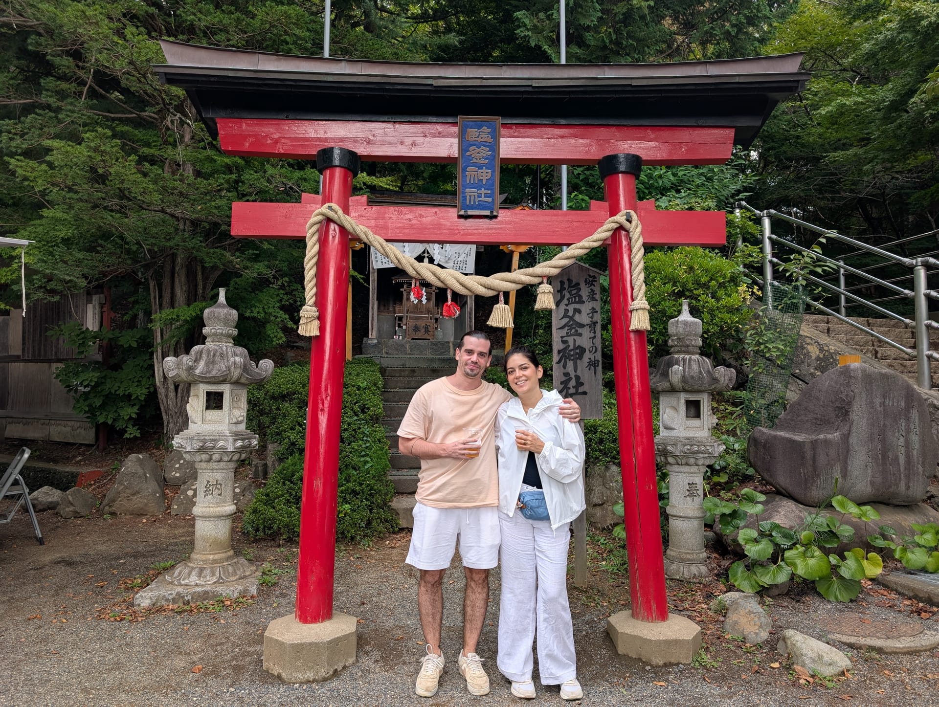 Pierre and Lia standing together under a red torii gate at the shrine, smiling, with stone lanterns and green trees behind them